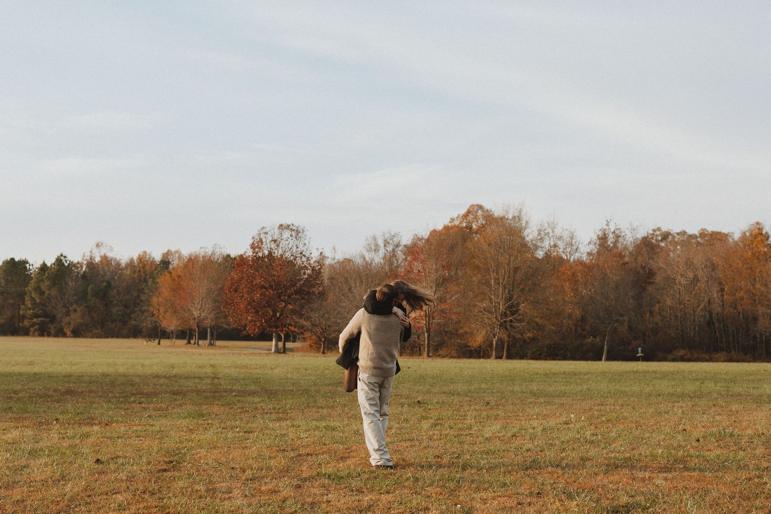 A person in a white sweater stands in an autumn field with orange trees in the background.