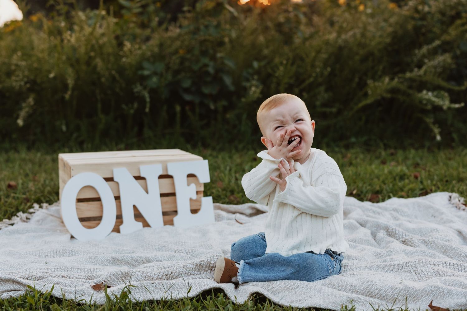 Baby in denim overalls crawls on white blanket with decorative letters spelling ONE in outdoor setting.