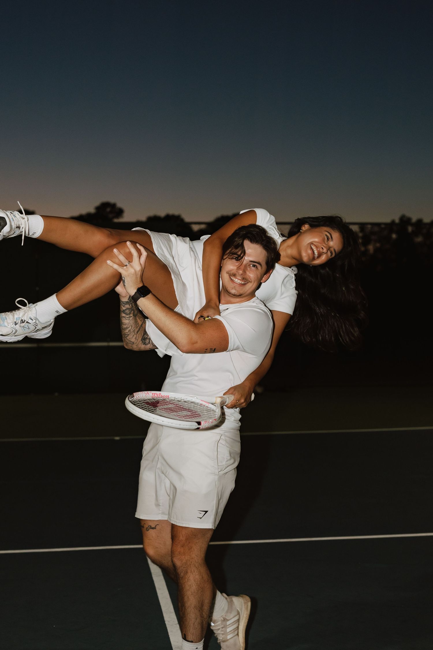 Two people in white outfits share a playful moment on a tennis court at sunset.
