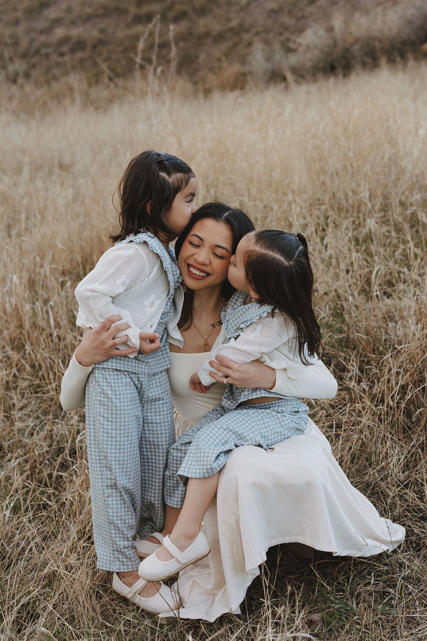 A mother sits in a golden field, smiling as her two young daughters in gingham outfits hug and kiss her cheeks.