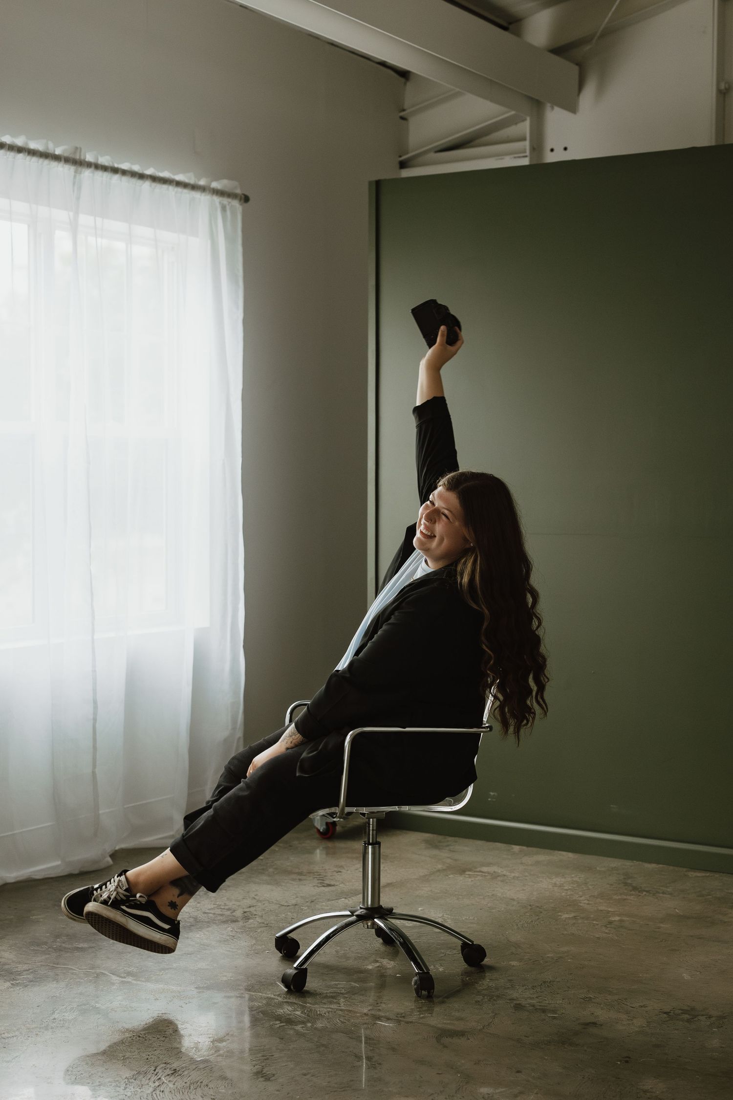 A person in black clothing falls backwards in an office chair in a minimalist room with white curtains.