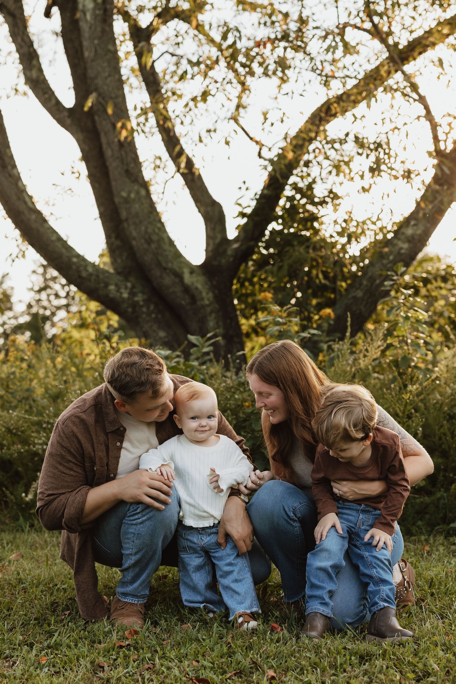 A family shares tender moments together while sitting on grass during golden hour sunset in a natural outdoor setting.