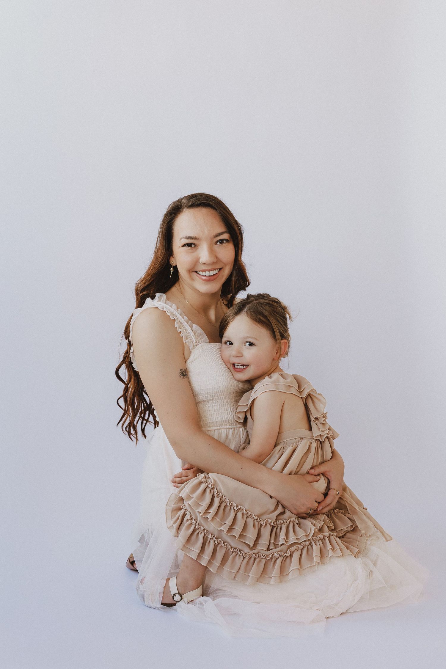 A smiling woman in a white dress hugs a young girl in a ruffled beige dress against a light background.