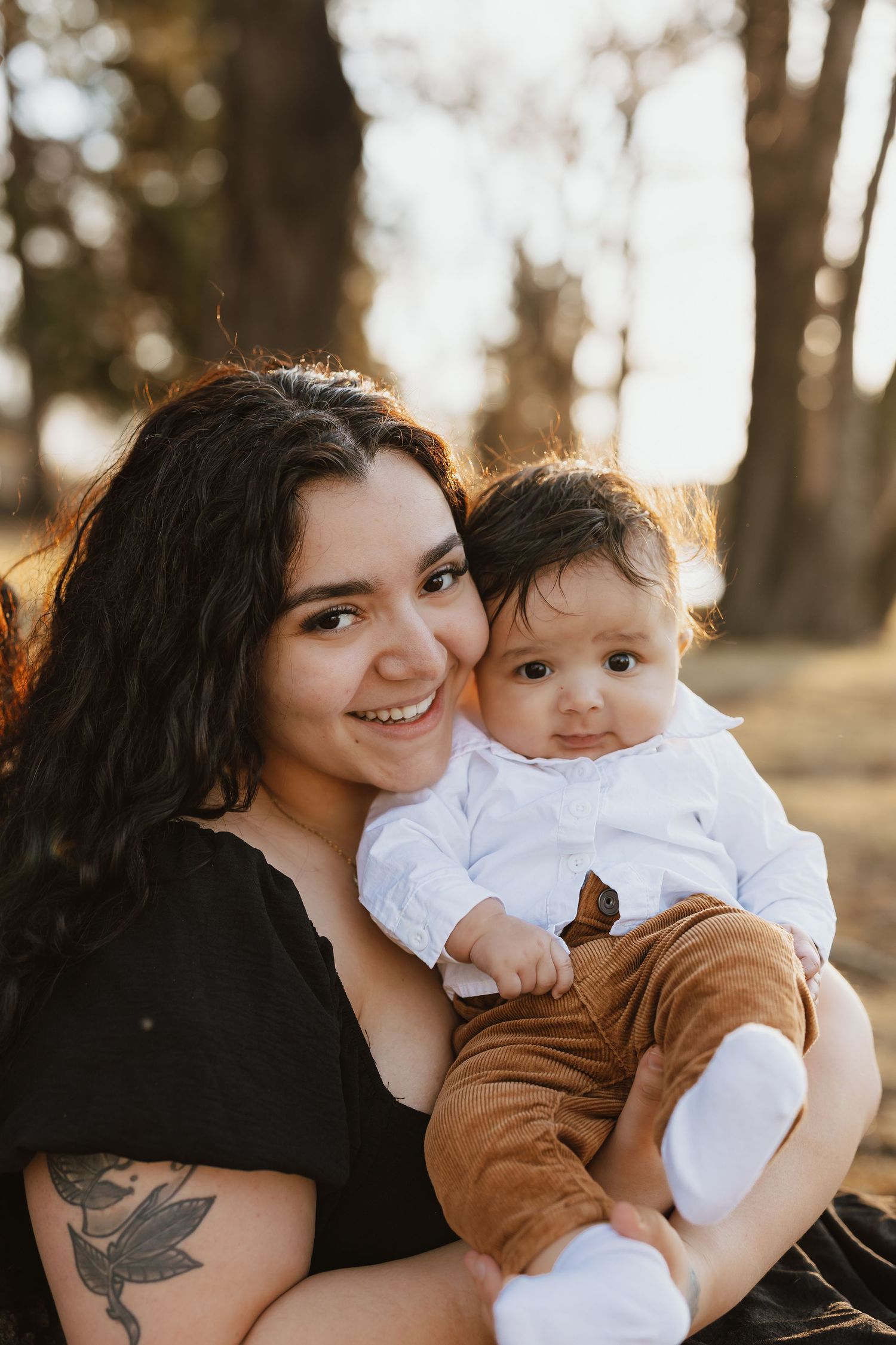 A smiling young woman with a tattoo holds a cute baby in a white shirt outdoors in a sunlit park.