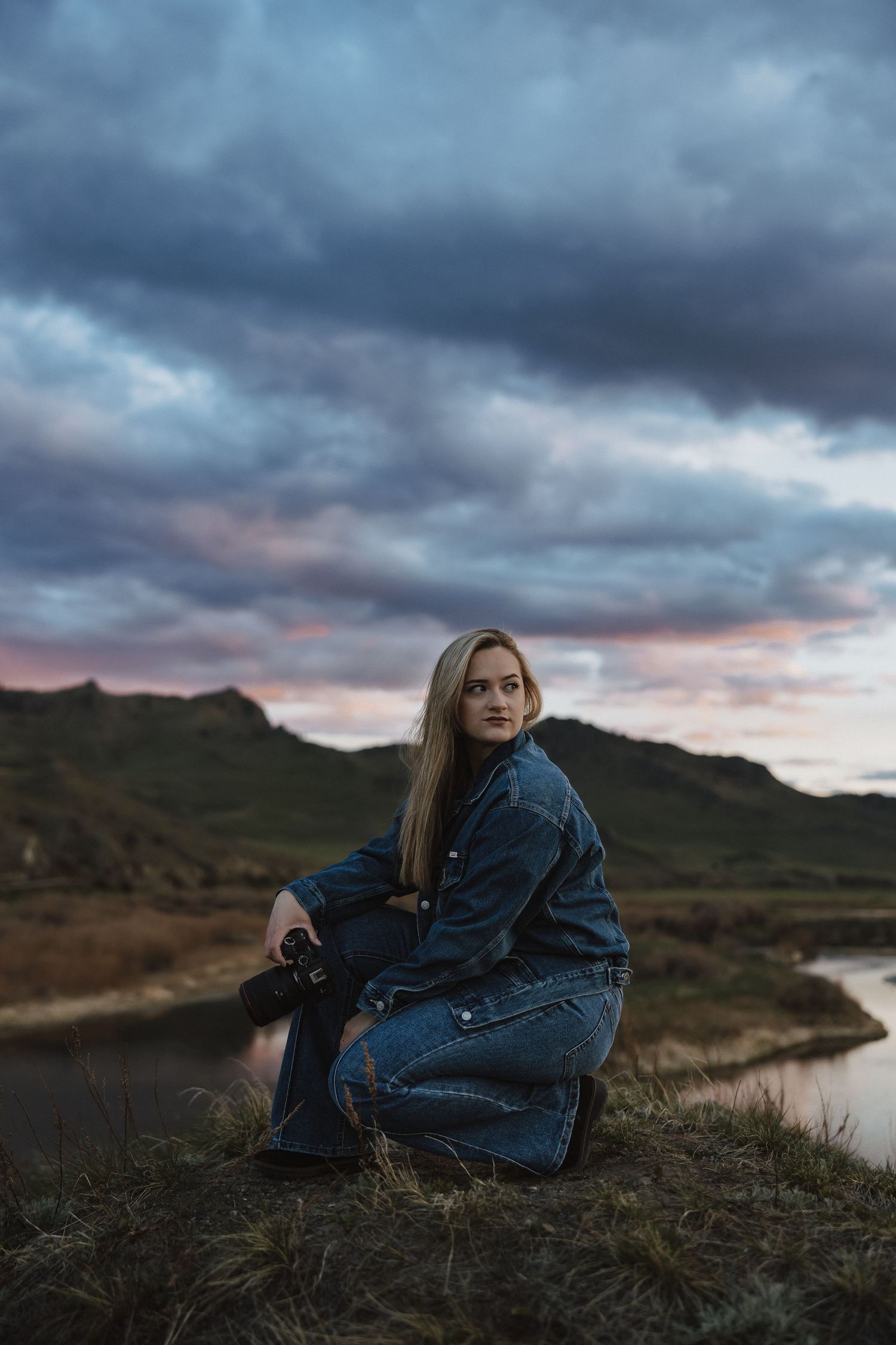 Woman in denim outfit sitting on hillside at dusk with dramatic cloudy sky and mountain lake backdrop.