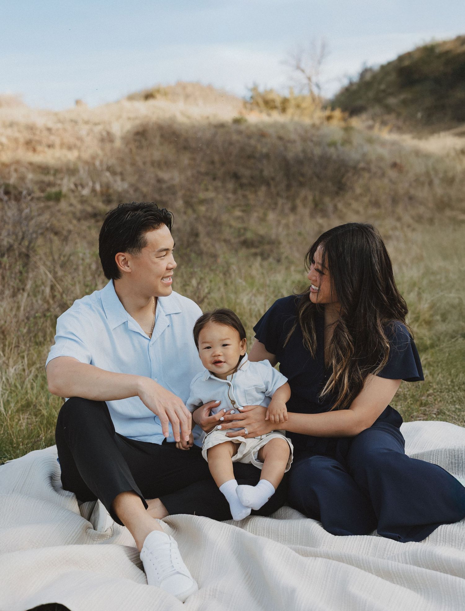 A young Asian family sits on a white blanket in a grassy field, parents smiling at their baby boy.