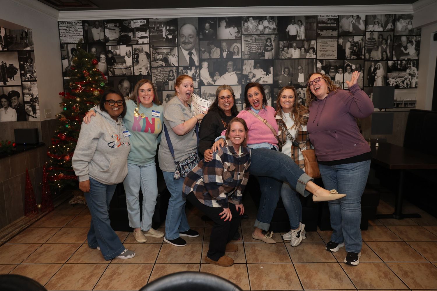 A group of people sitting together in a casual indoor setting with wood floors and holiday decorations visible.