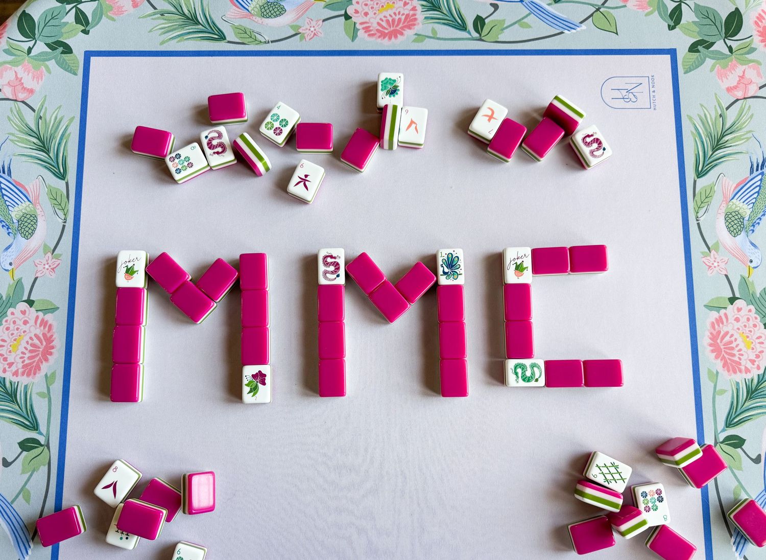 Pink wooden blocks and letter dice arranged to spell MME on a floral patterned background with scattered pieces.