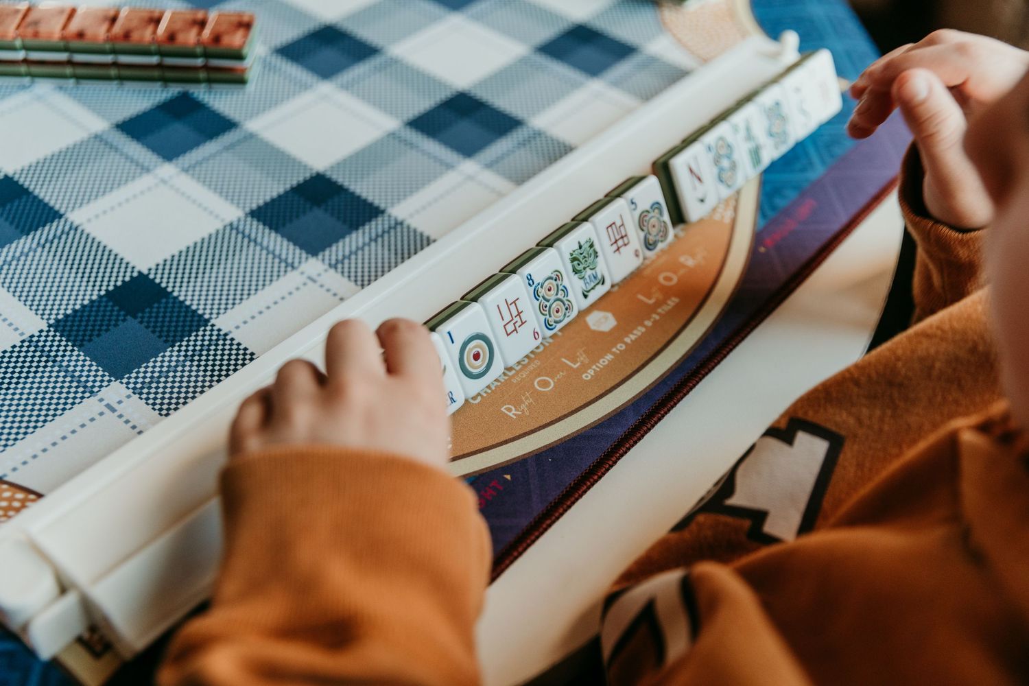 Close-up of hands playing mahjong on a checkered tablecloth with white game tiles.
