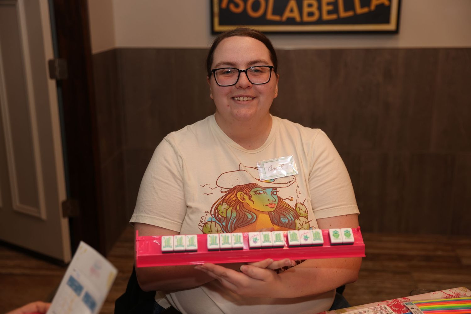 A person smiles while sitting at a table with Scrabble letter tiles and game pieces arranged in front of them.