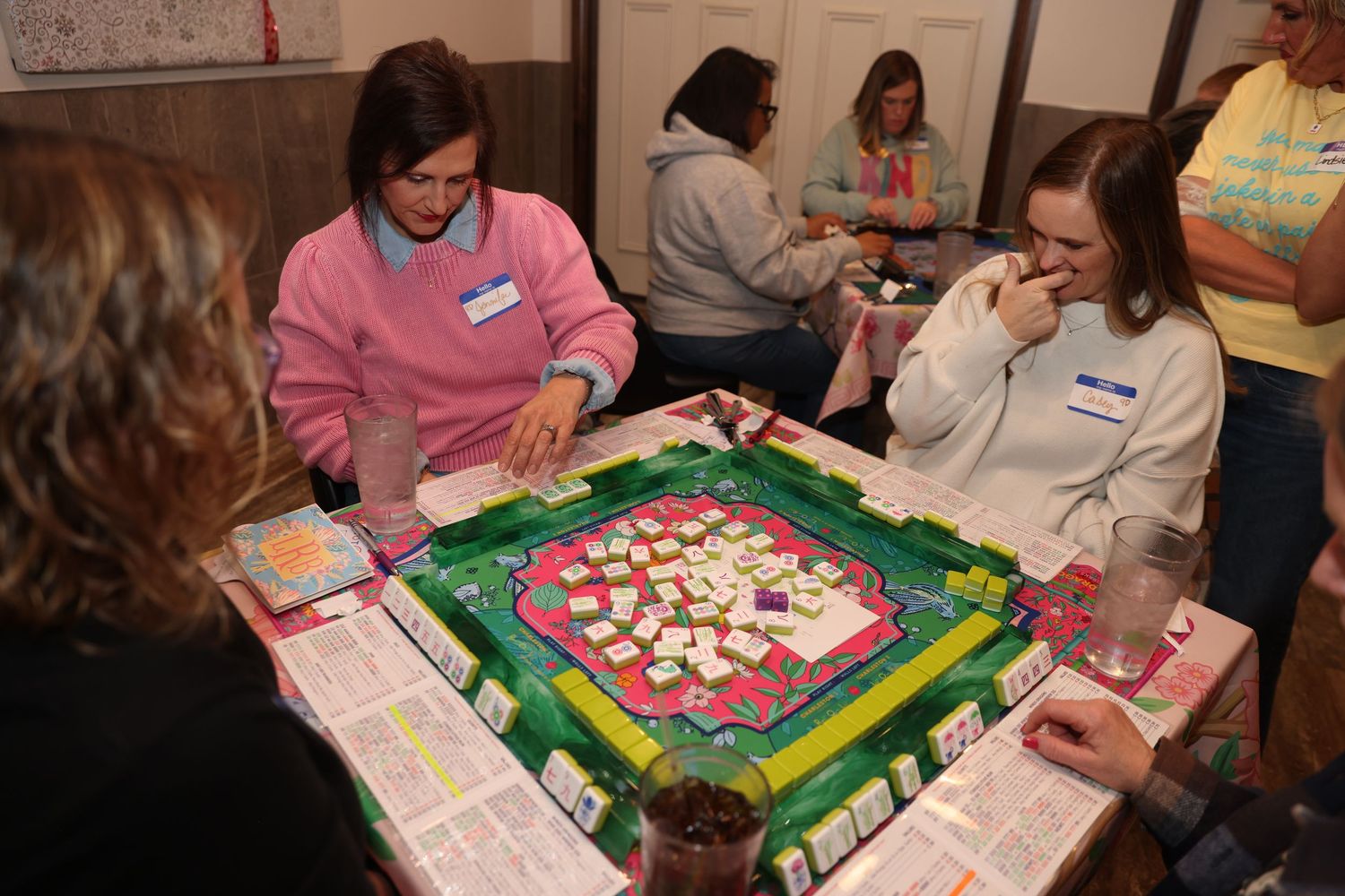 People gathered around a table playing mahjong with pink and white tiles on a green felt surface.