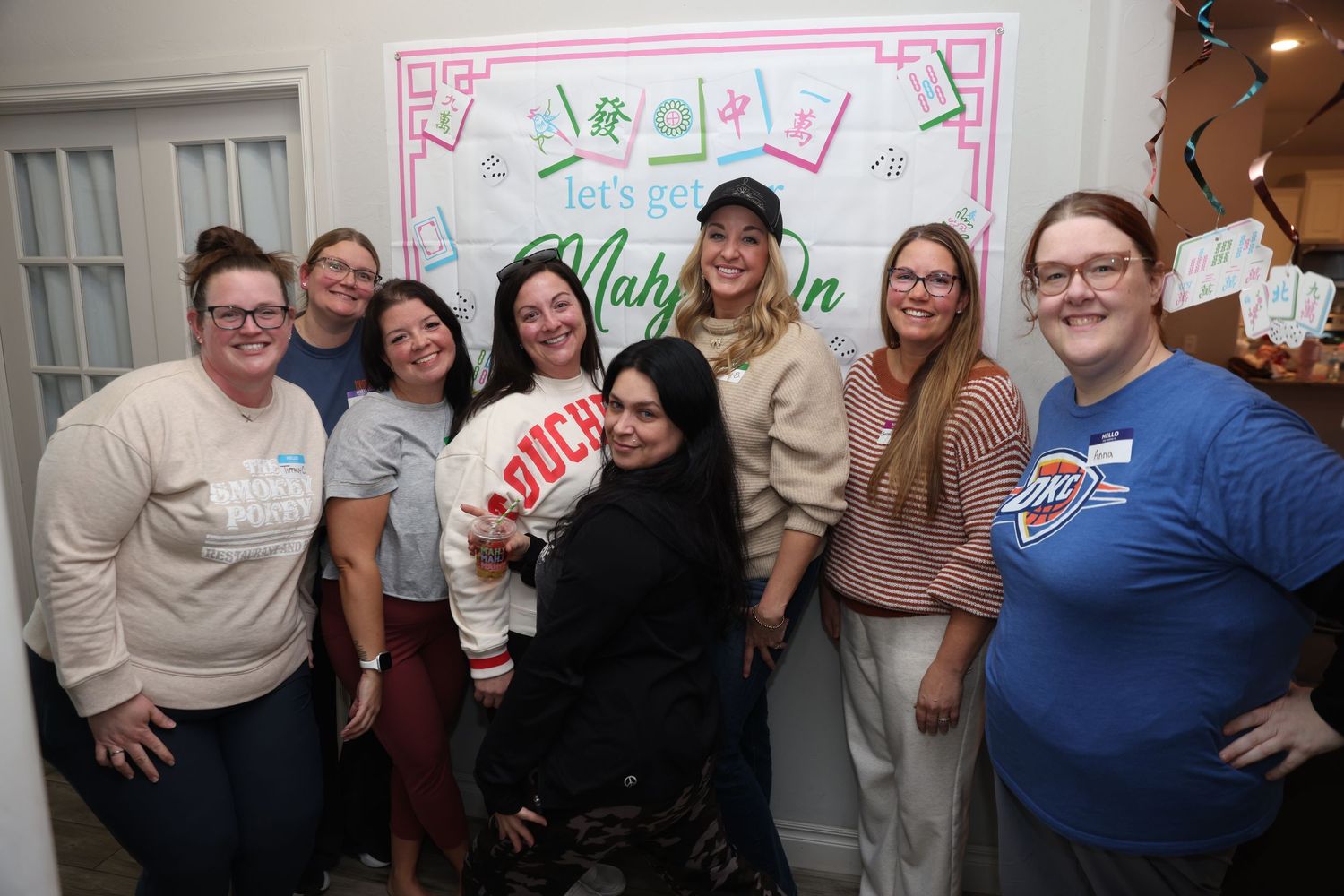 A group of people celebrate at a party with pink decorative elements on the wall behind them.
