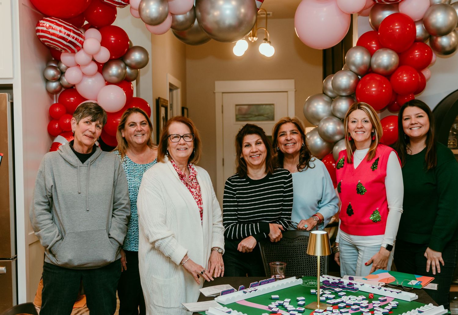A group gathers around a decorated table with red and silver balloons for a festive celebration.