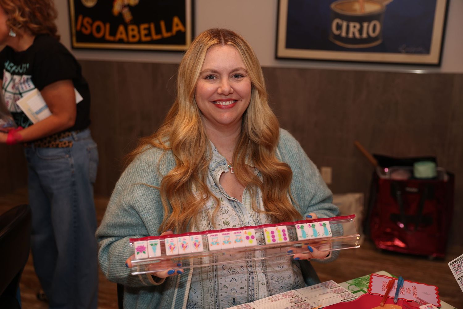 Someone smiles while holding a row of wrapped holiday gifts at a casual indoor gathering with vintage beer signs on wall.