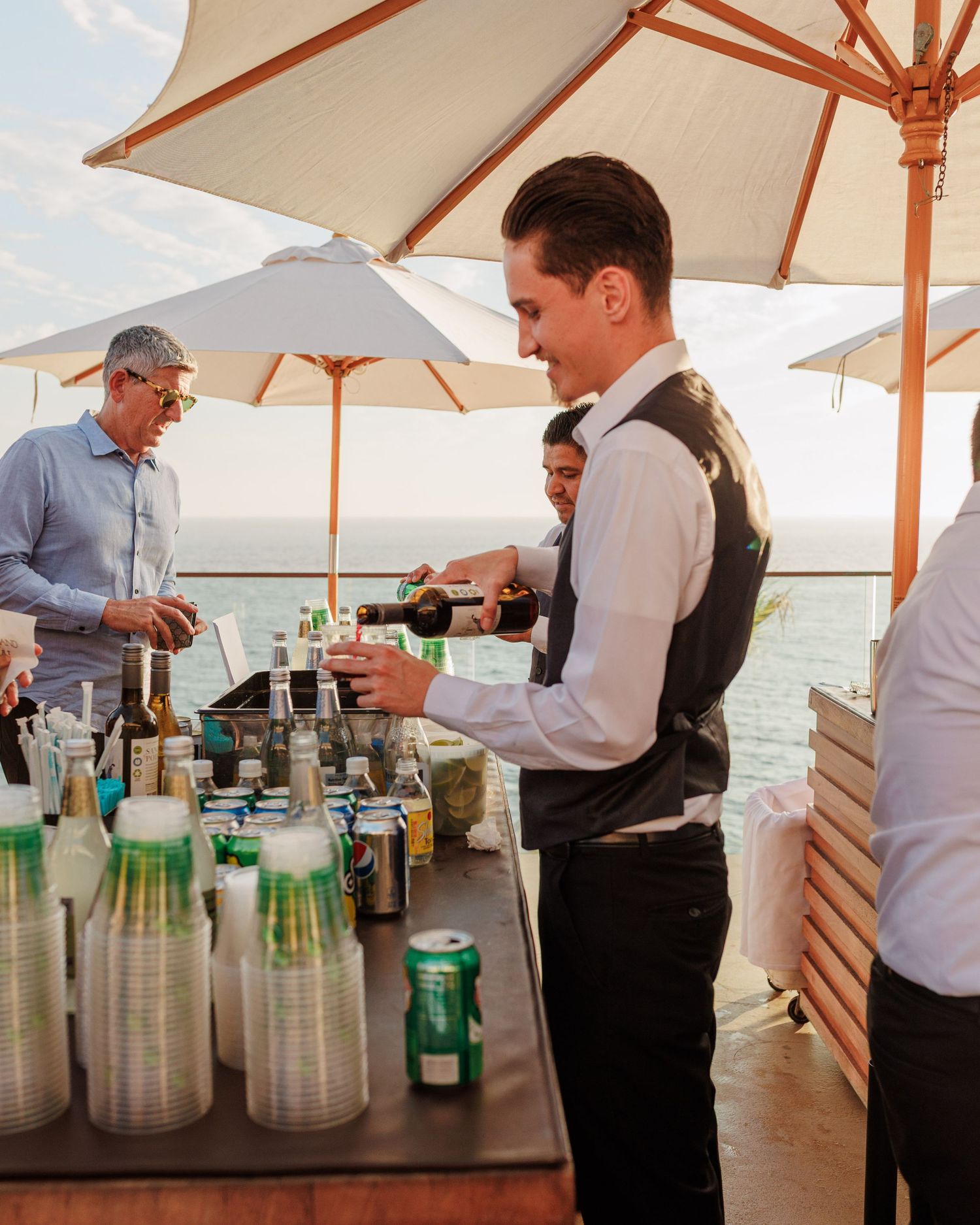 Bartender pouring drinks at an outdoor wedding bar with ocean views and white umbrellas.