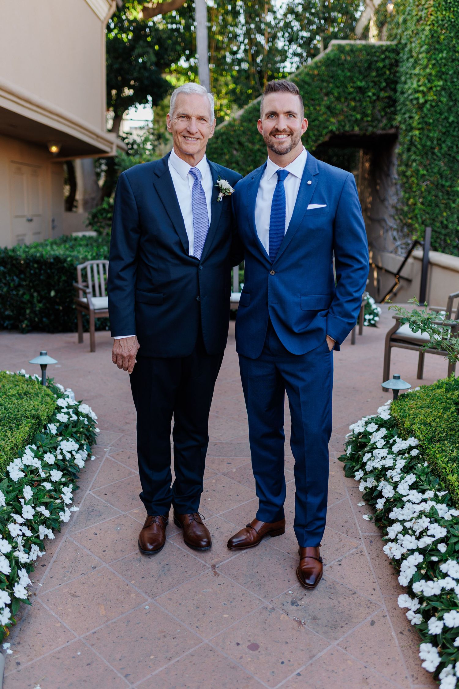 Groom in navy blue suit poses with father in dark suit amid white flowers at wedding venue.