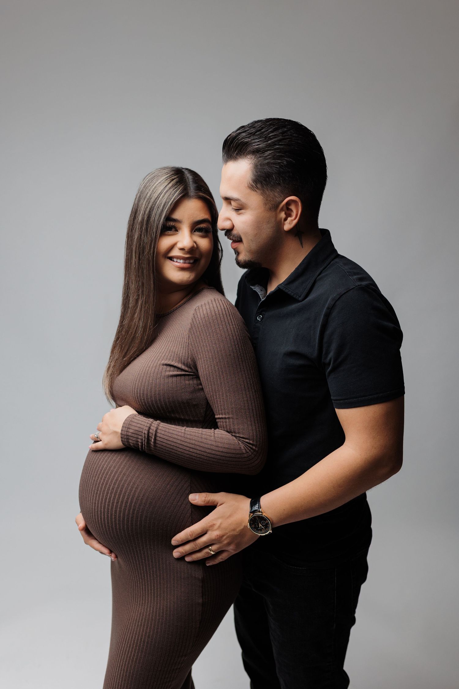 Intimate maternity portrait featuring couple in dark tones against light background.