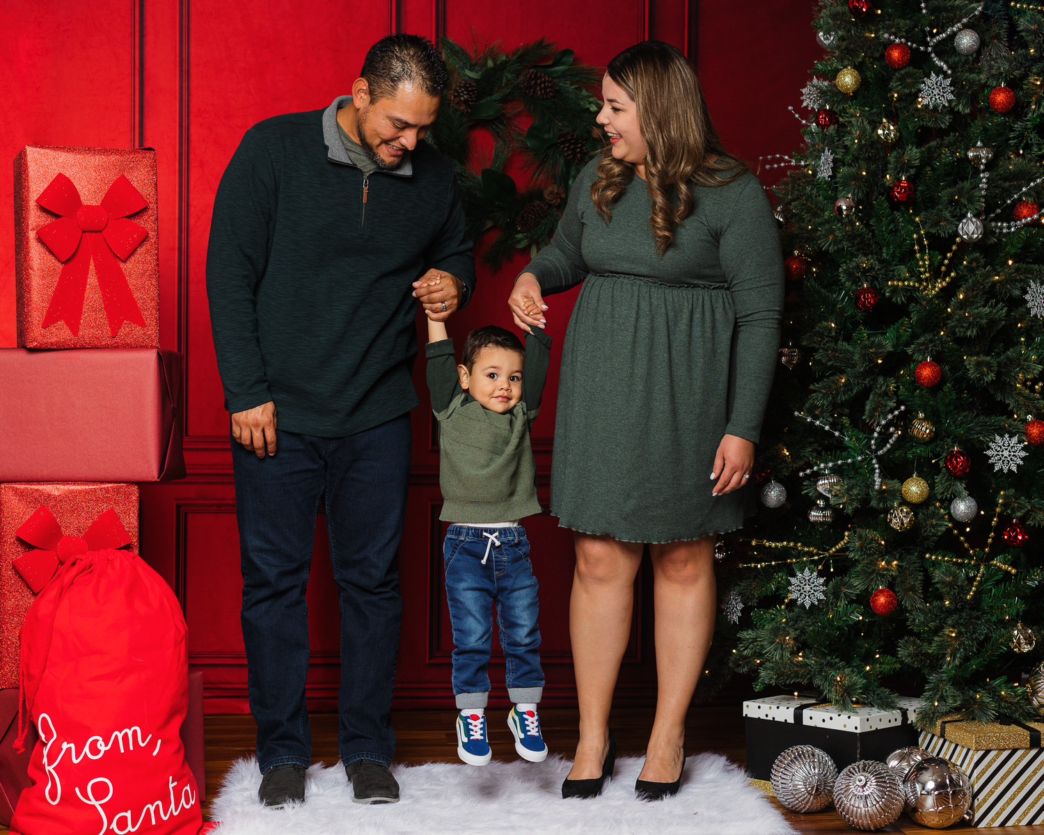 Family of three posing by decorated Christmas tree with gifts, parents holding toddler son hands in holiday outfits.