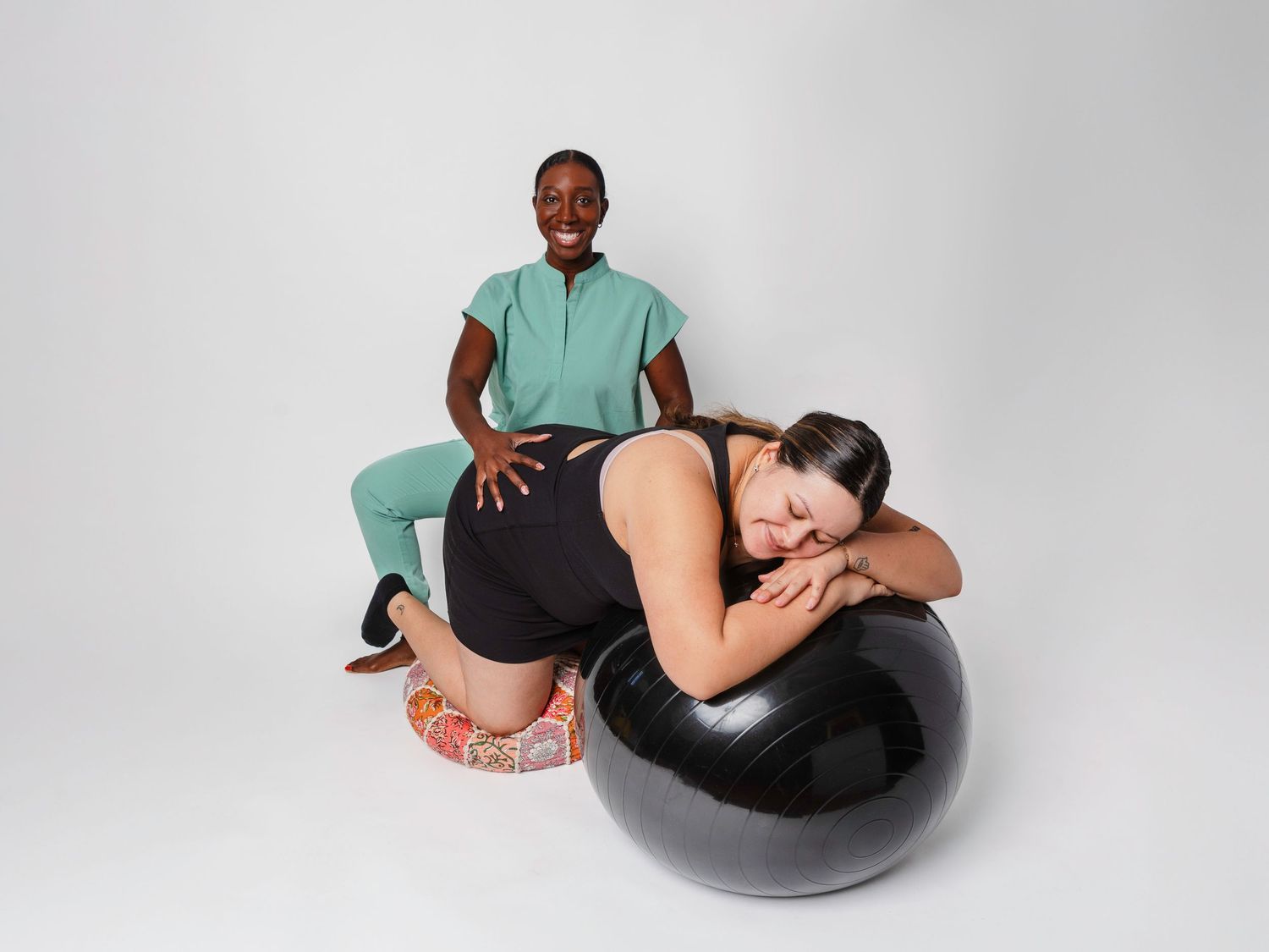 A doula in green scrubs supports a pregnant woman resting on a black exercise ball during labor preparation.