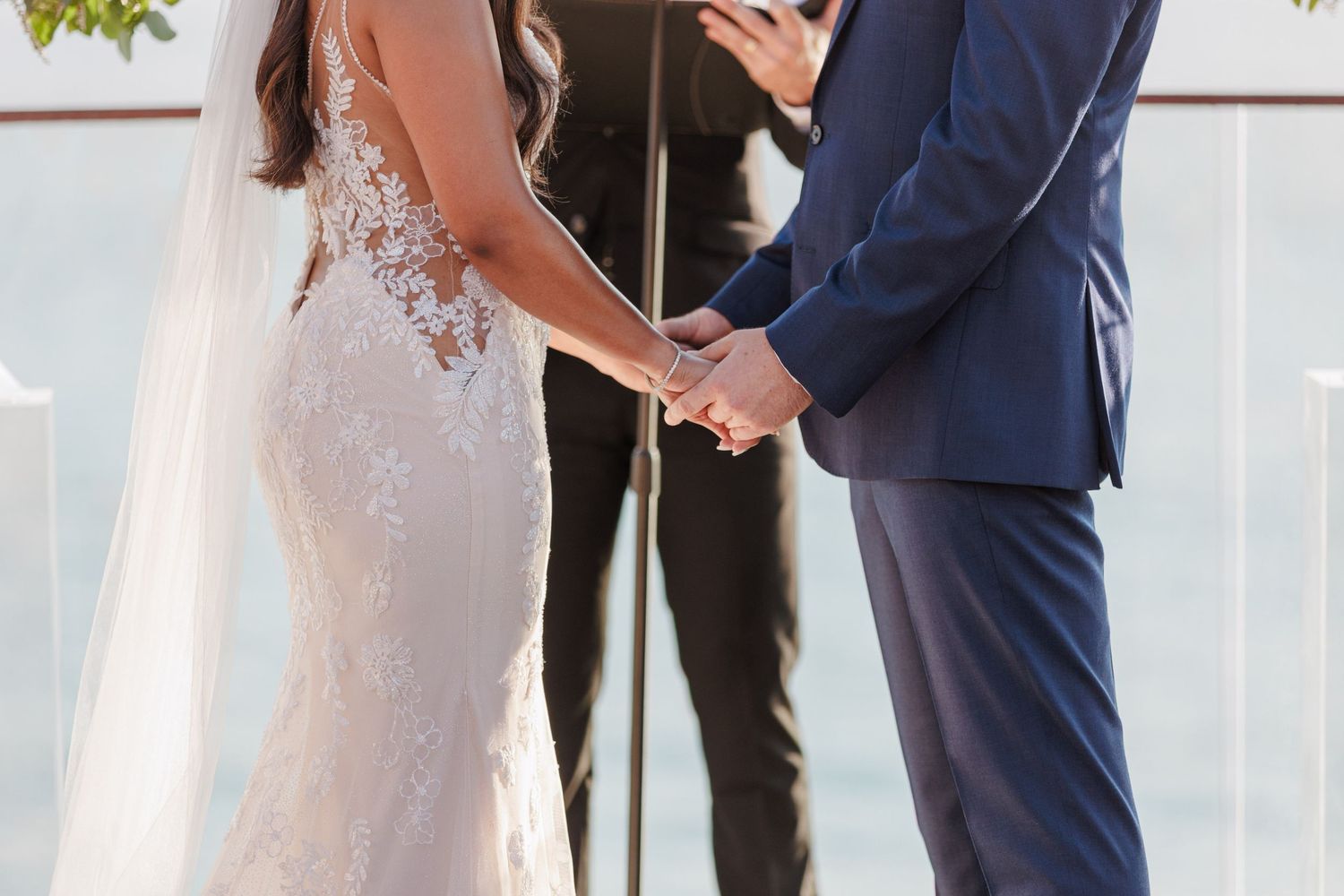 Bride and groom hold hands during outdoor wedding ceremony, bride wearing lace mermaid gown with veil.