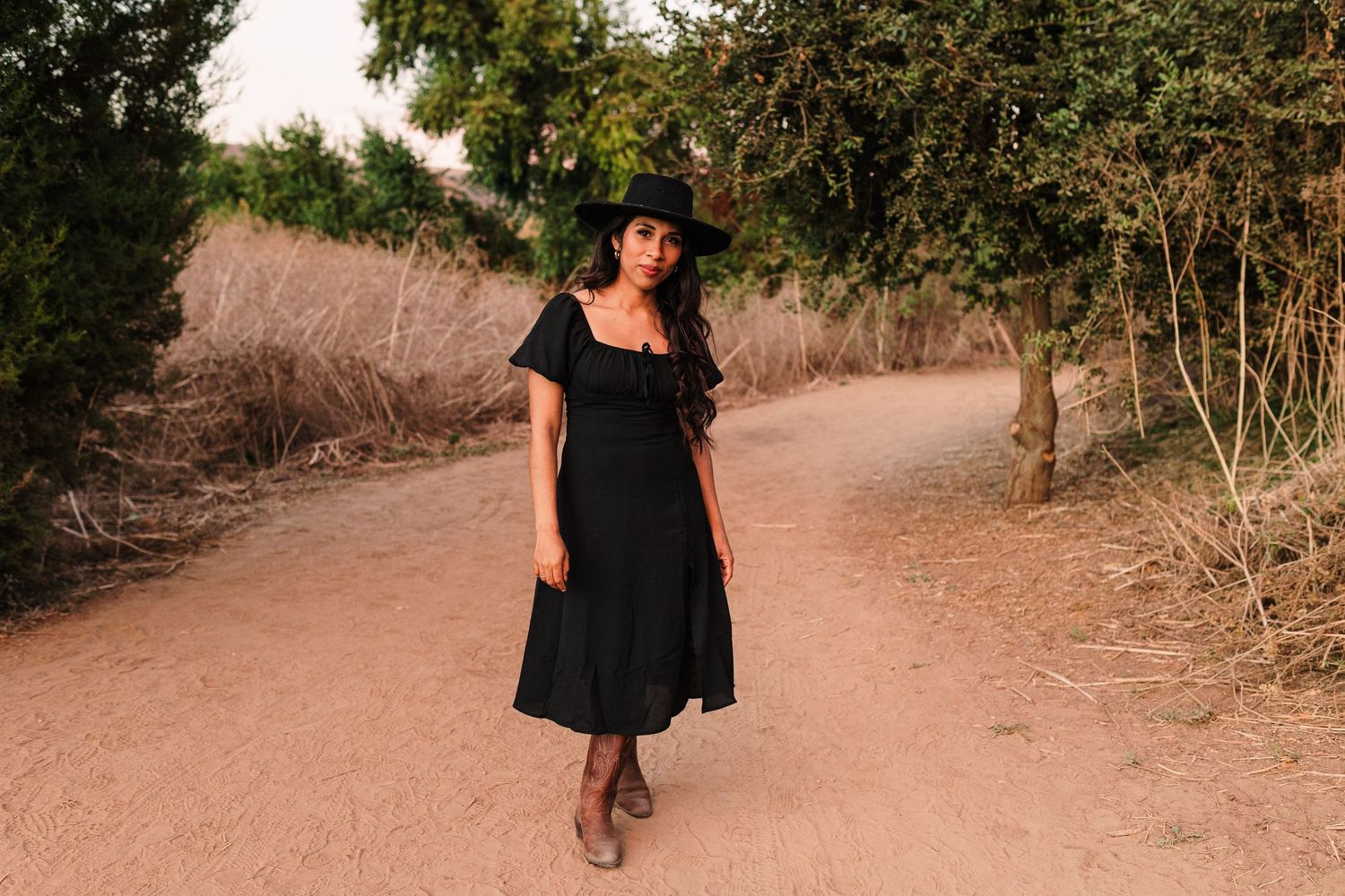 A person in a black dress walks along a desert path during sunset.