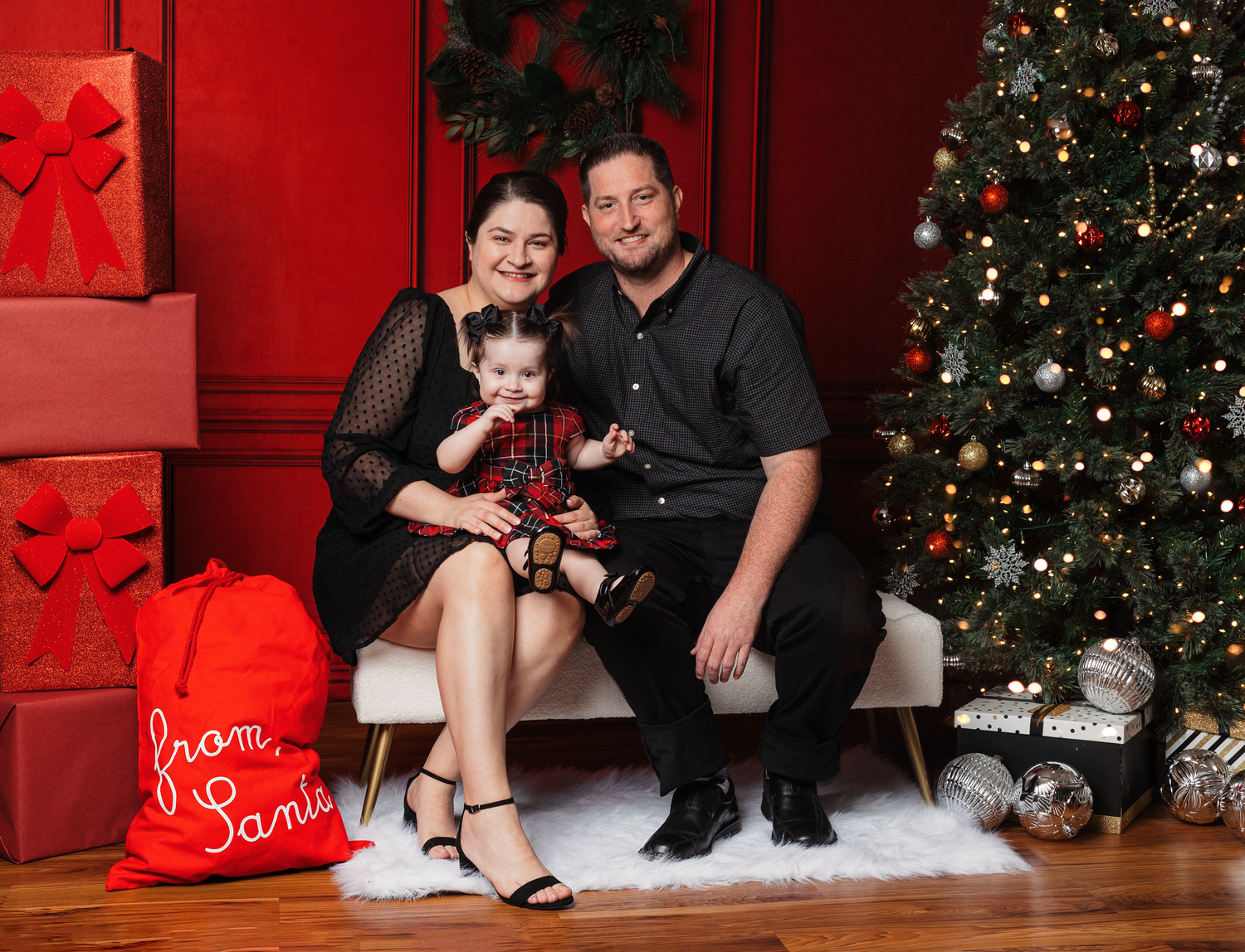 Christmas family photo setup with decorated tree, white bench, and red wall background.