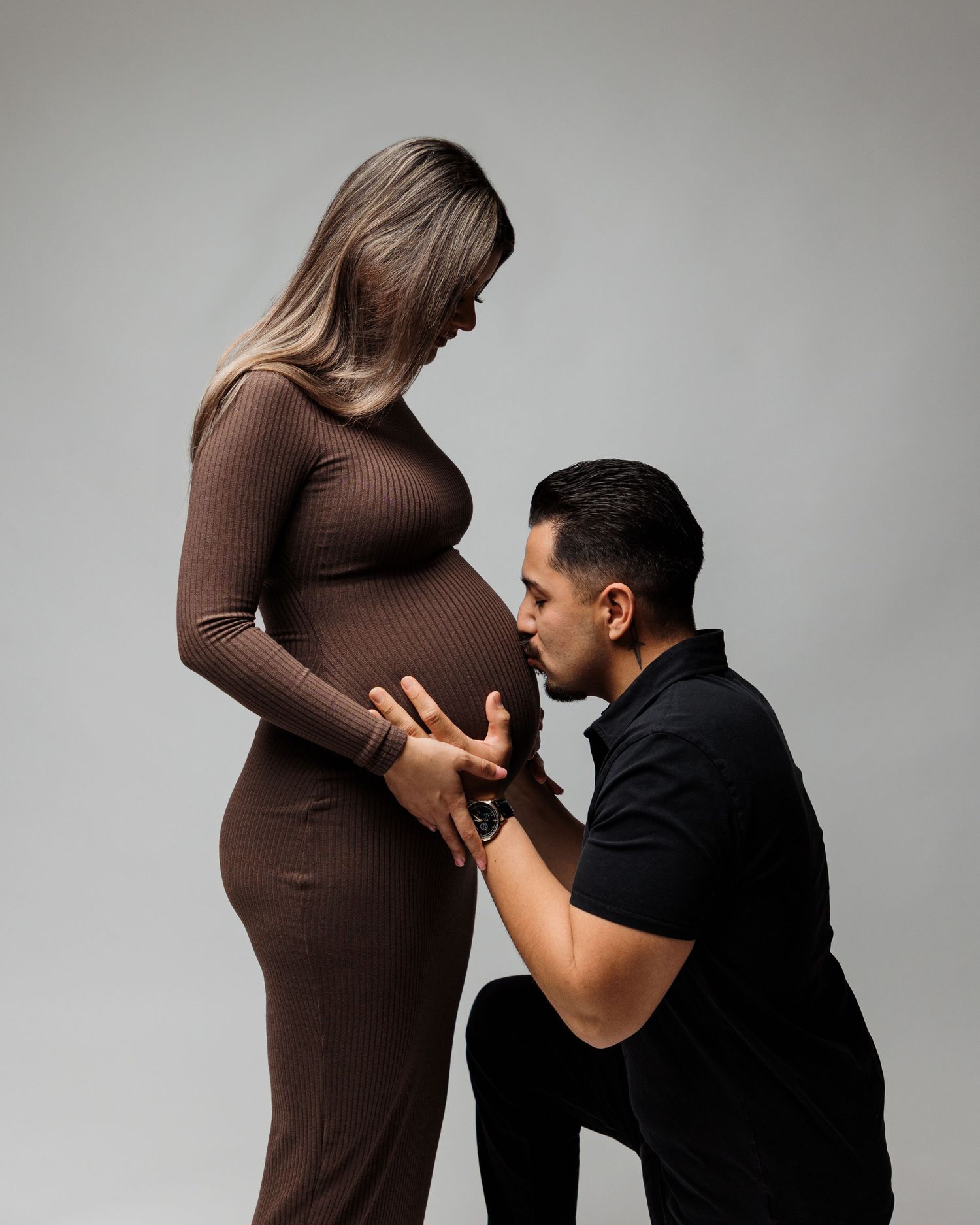 Tender moment between couple during pregnancy photoshoot with brown dress against grey background.