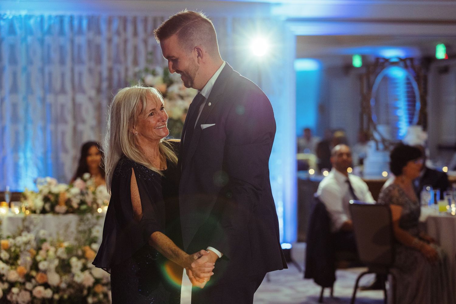 Tall groom shares a tender mother-son dance moment at elegant wedding reception with blue lighting.