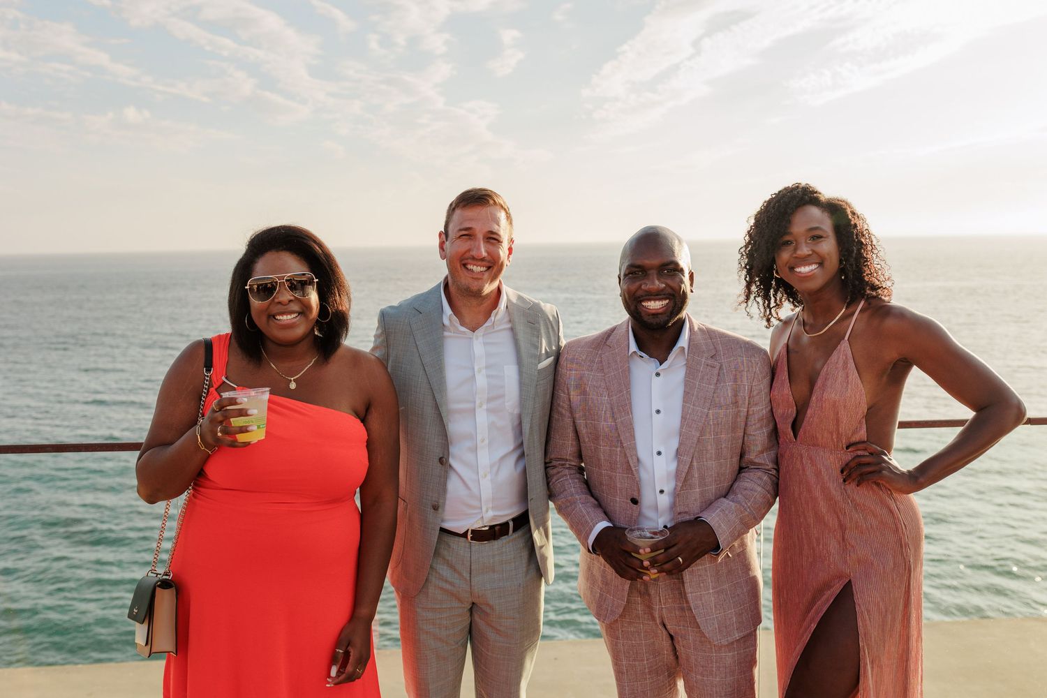 Four guests smile together on an oceanfront terrace at a sunset wedding celebration.