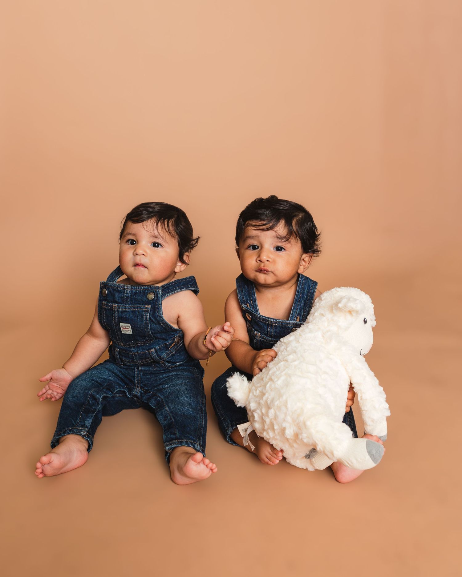 Twin toddlers in denim overalls sit together with white plush toy on peach background.