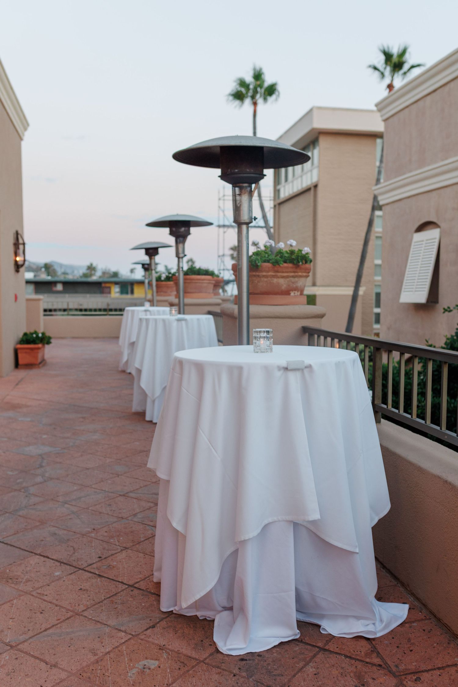 Outdoor cocktail hour setup with white draped high-top tables and standing patio heaters on terrace.