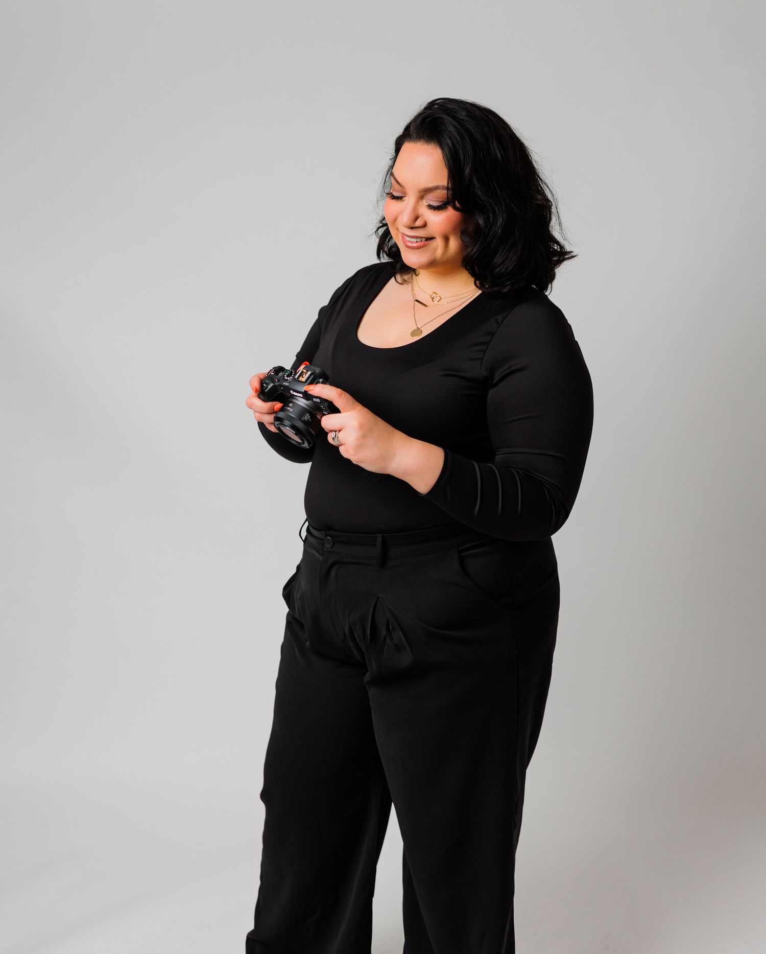 A woman in all-black outfit holds a camera, smiling downward against a light gray studio background.