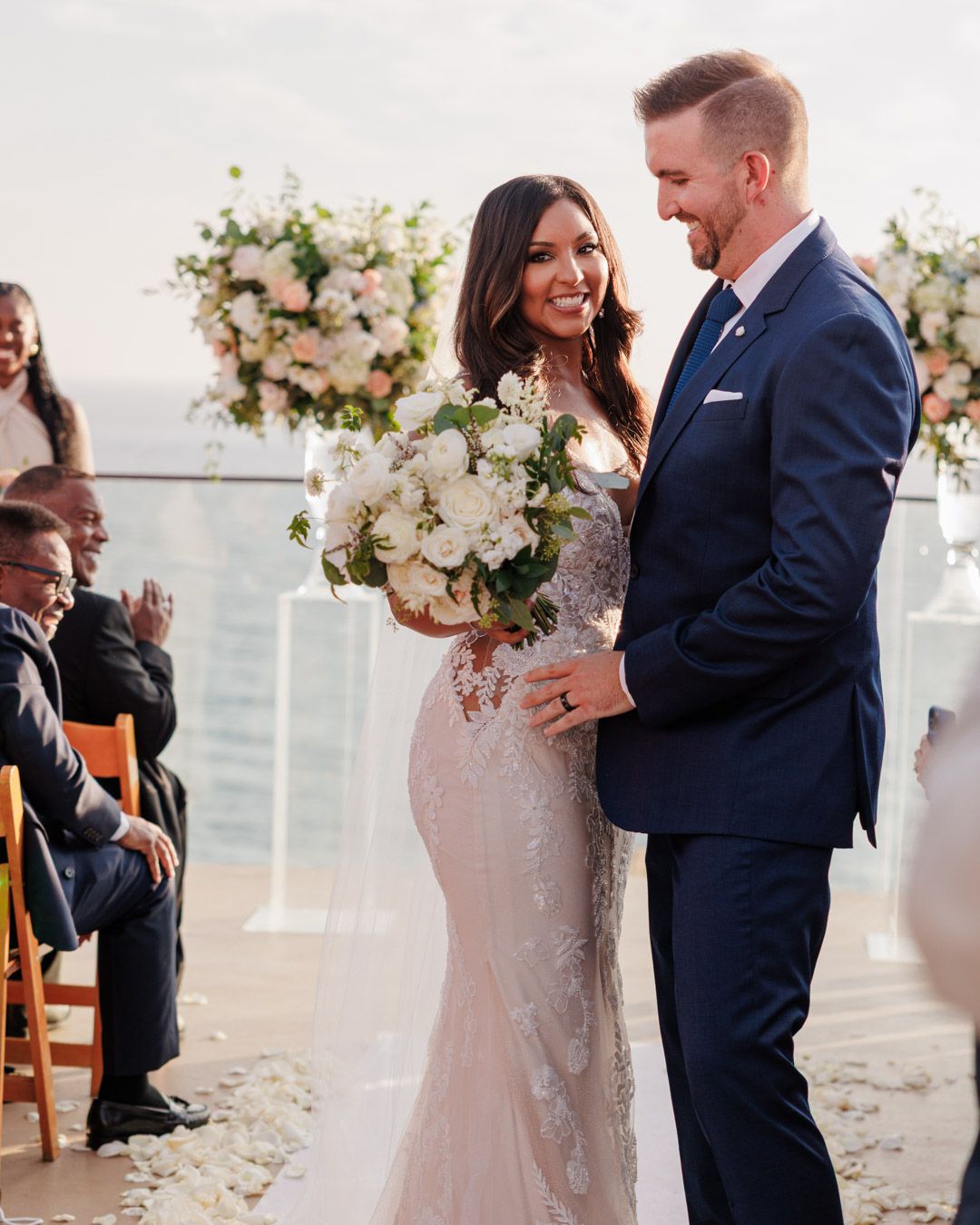 Bride in lace gown holding white bouquet stands with groom in navy suit during outdoor wedding ceremony by the ocean.