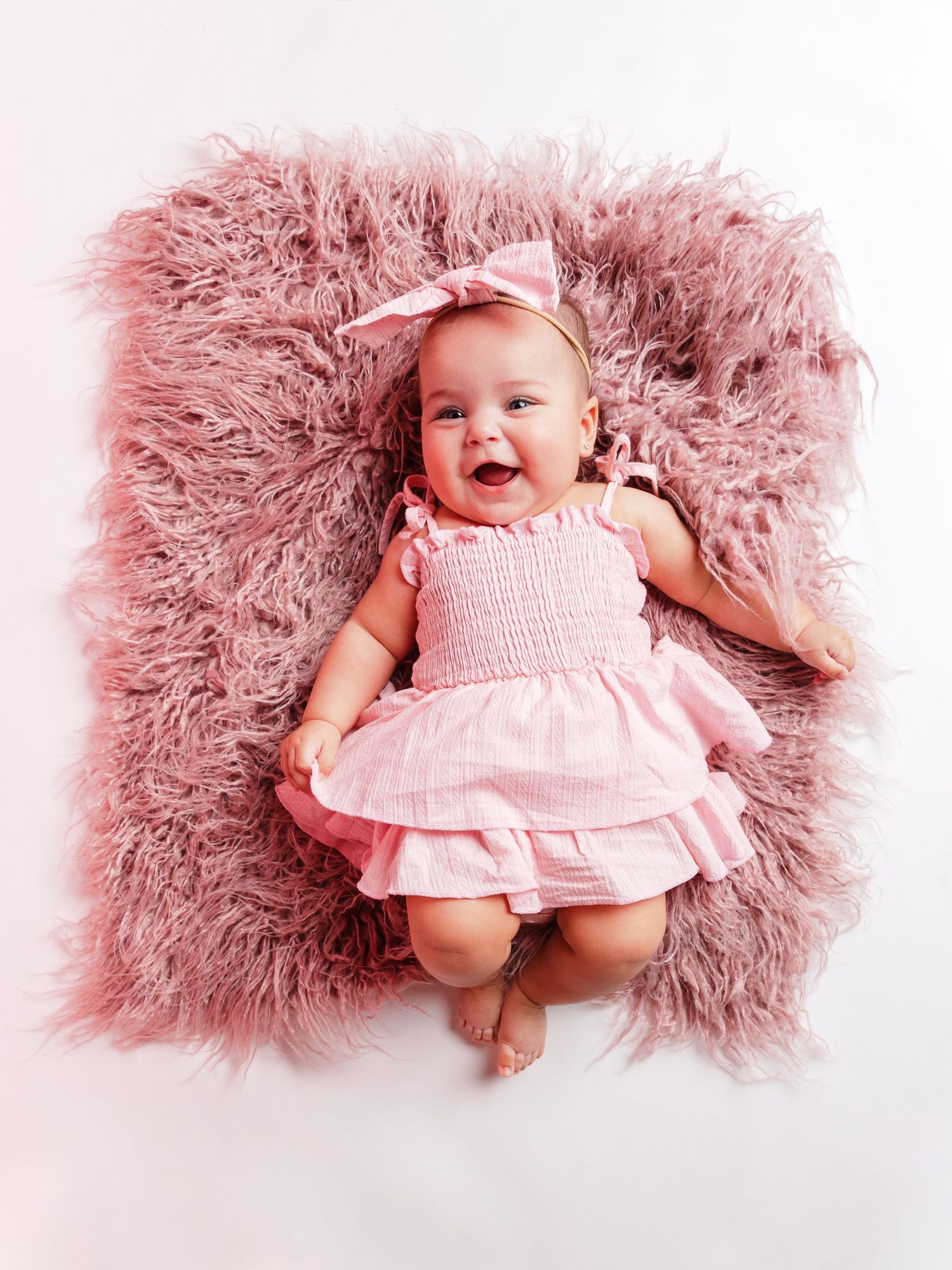 Smiling baby girl in pink dress and bow headband lying on fluffy pink rug.