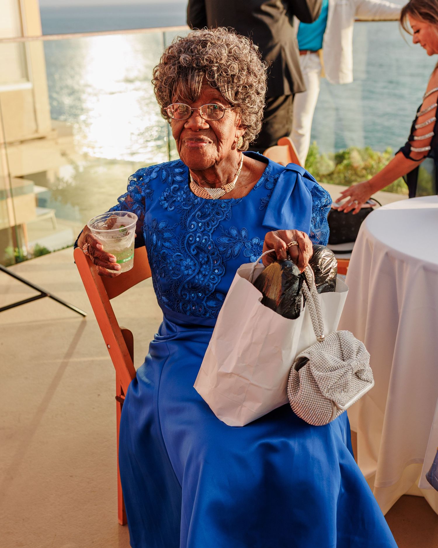Elderly woman in blue dress holding a bag and drink at an outdoor wedding reception by the ocean.