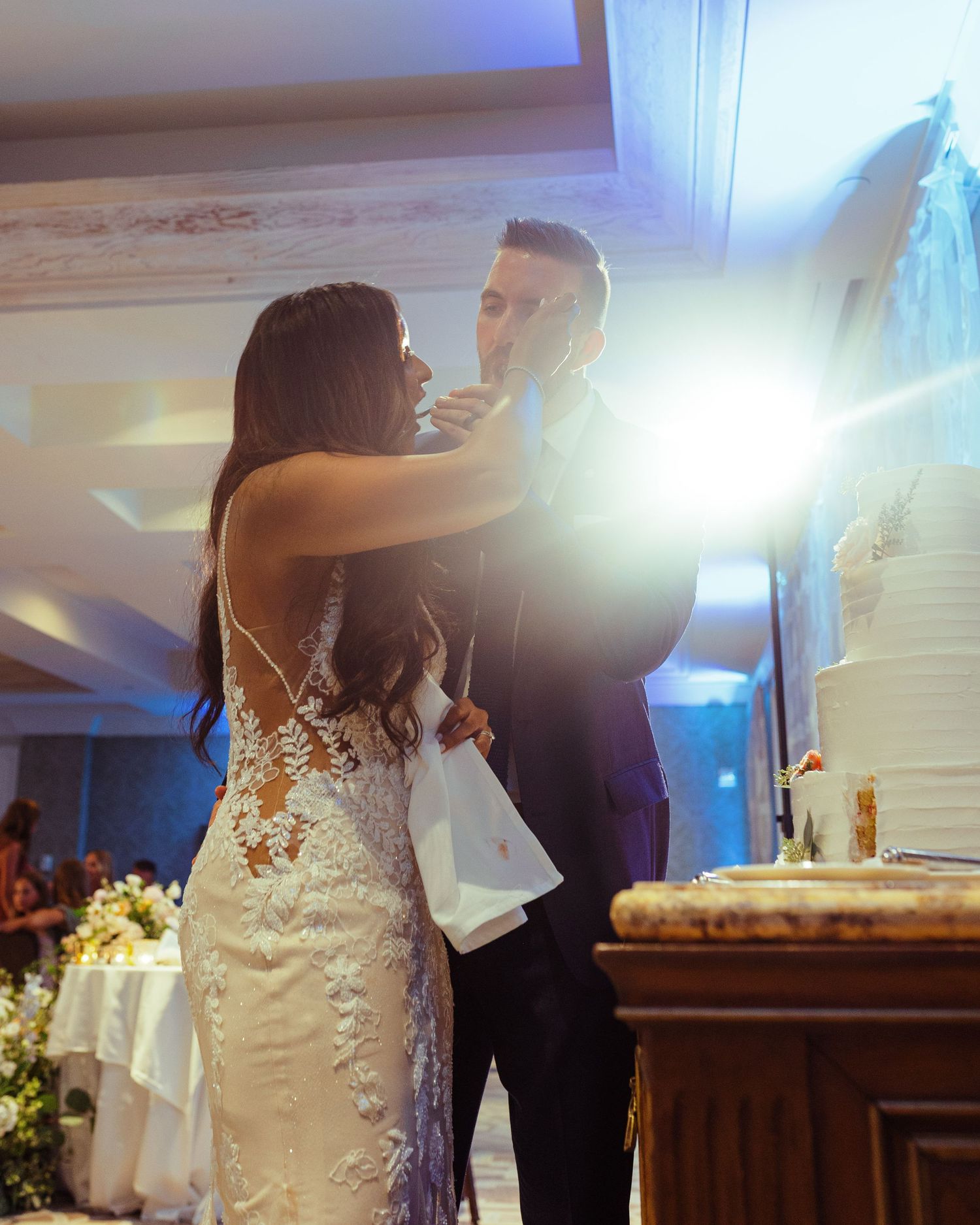 Bride feeding wedding cake to groom in elegant lace dress at romantic reception with white tiered cake.