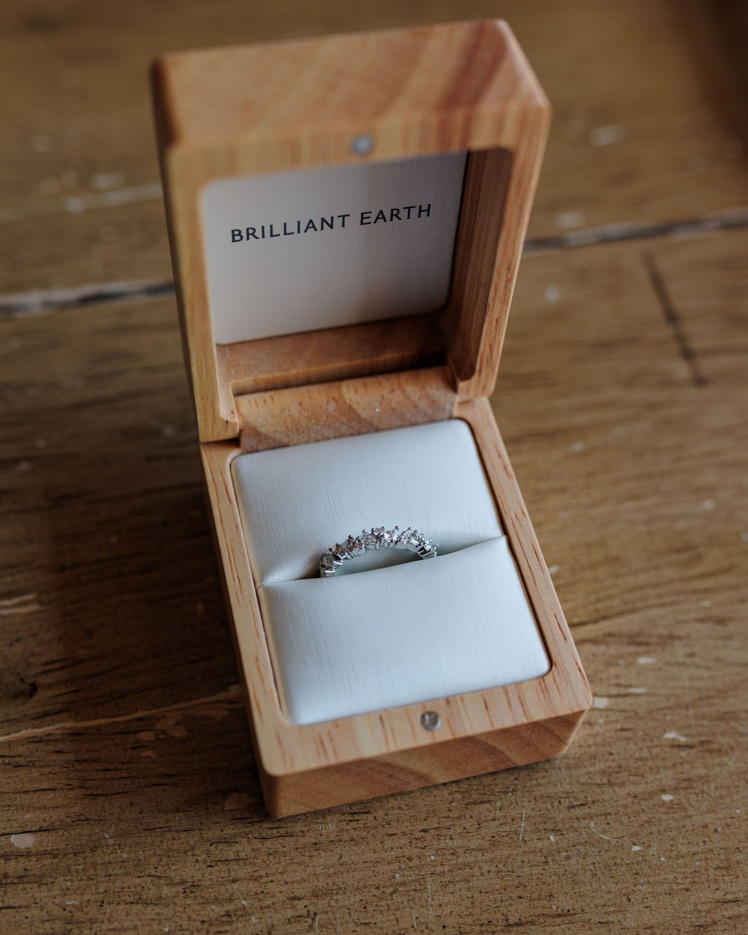 Brilliant Earth diamond engagement ring displayed in a wooden ring box on a rustic wooden surface.