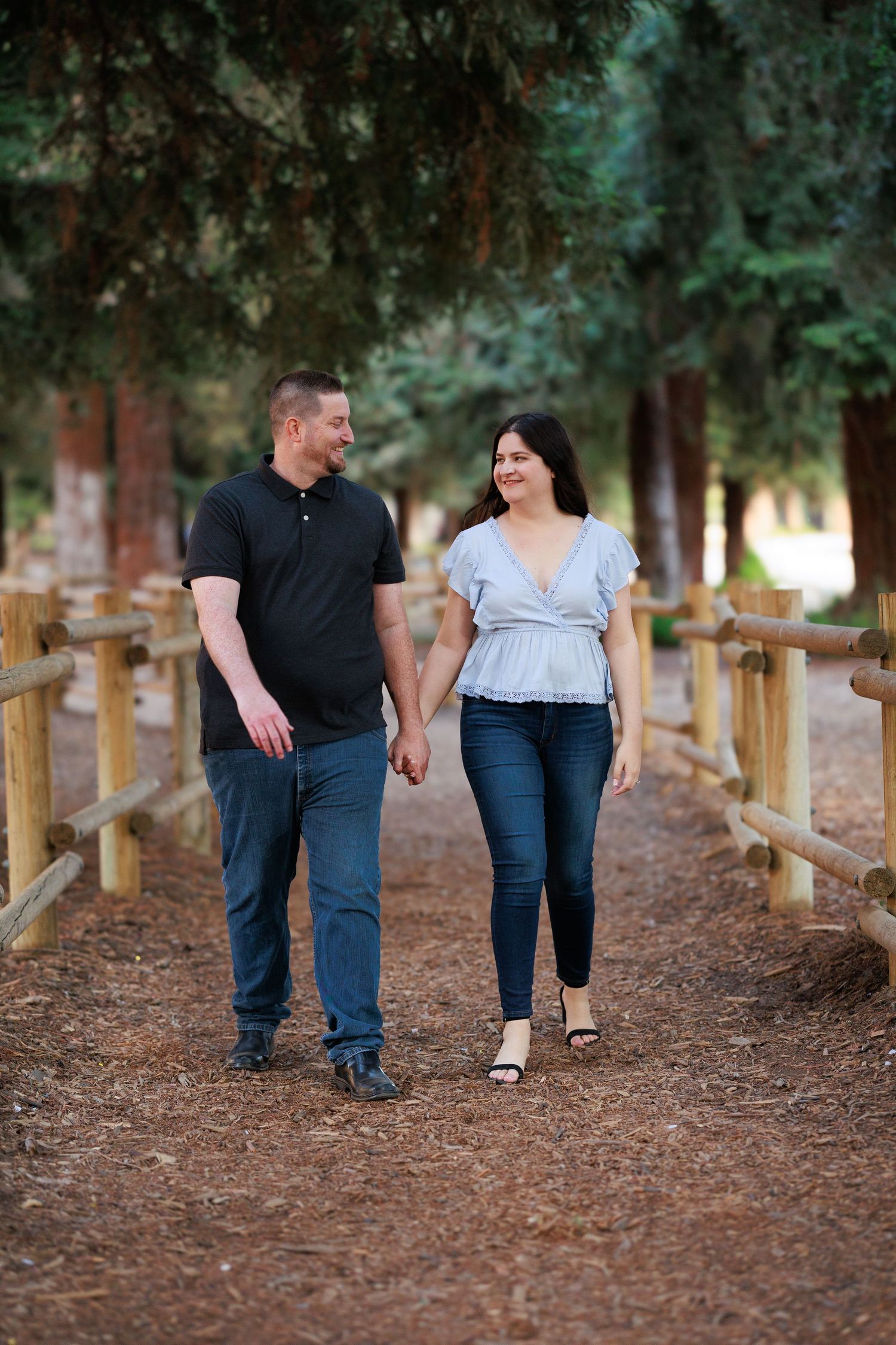 A couple walking hand in hand along a rustic wooden fence path surrounded by tall evergreen trees.