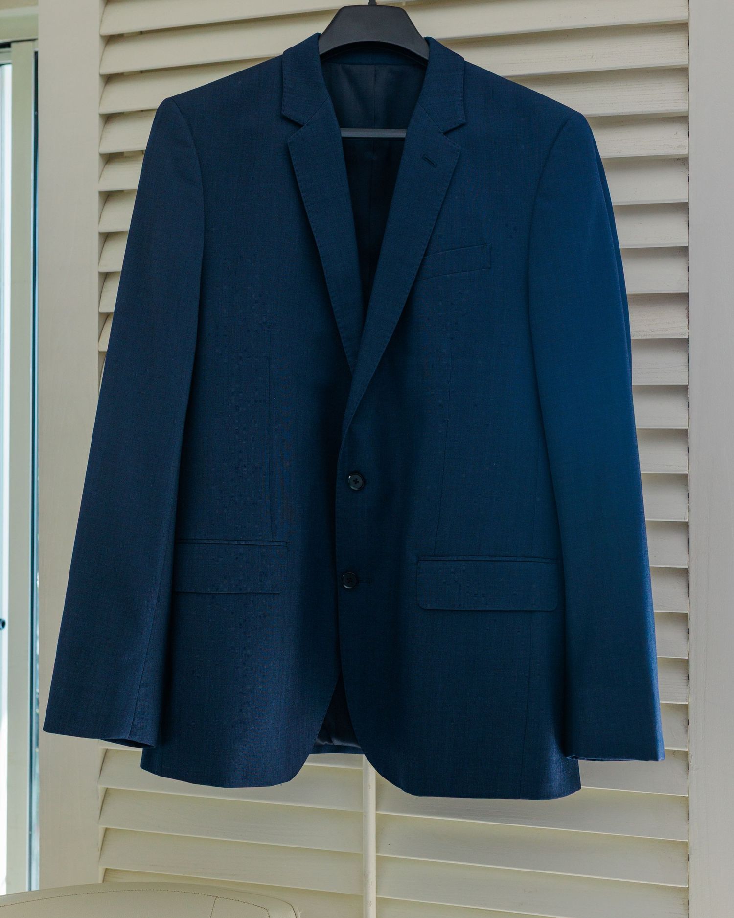 Navy blue two-button men's suit jacket on hanger against white shuttered window.
