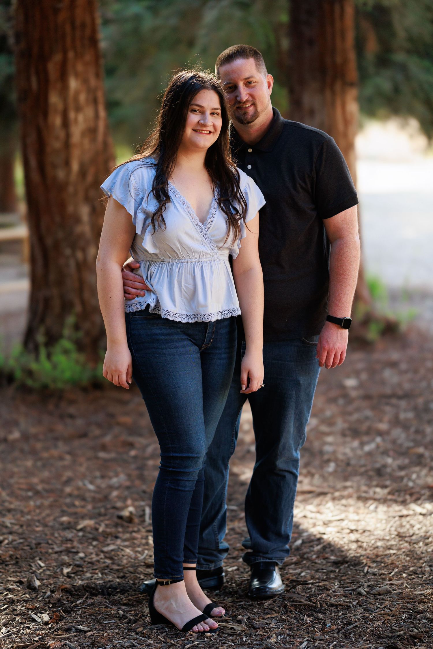 Couple in casual attire standing together on pine needle covered ground in a forest.