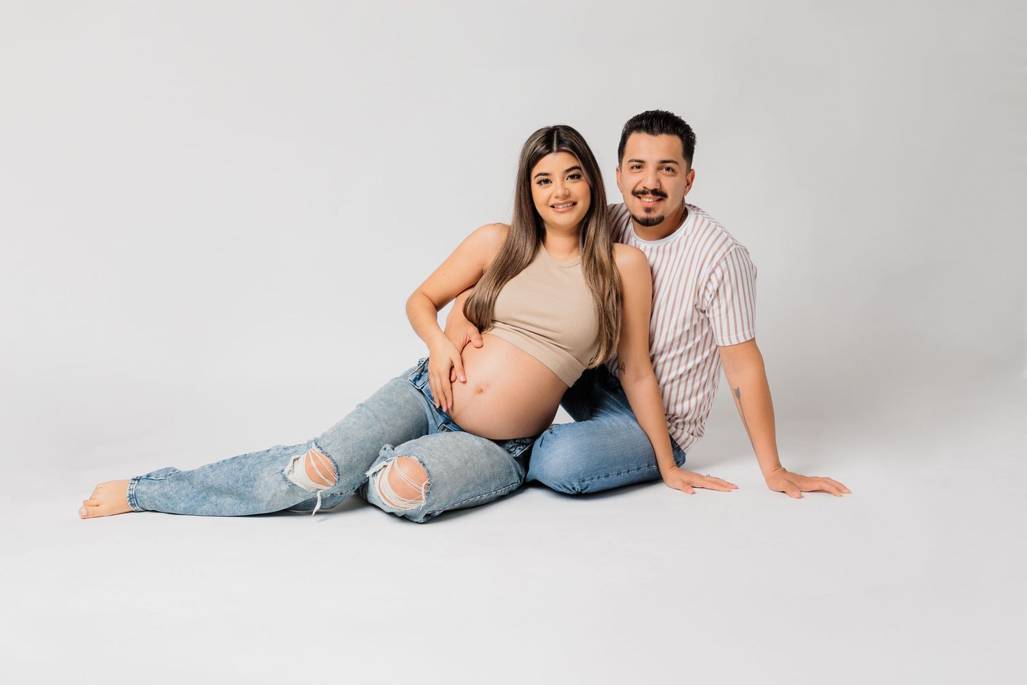 Relaxed maternity portrait with couple in matching casual outfits on white backdrop.