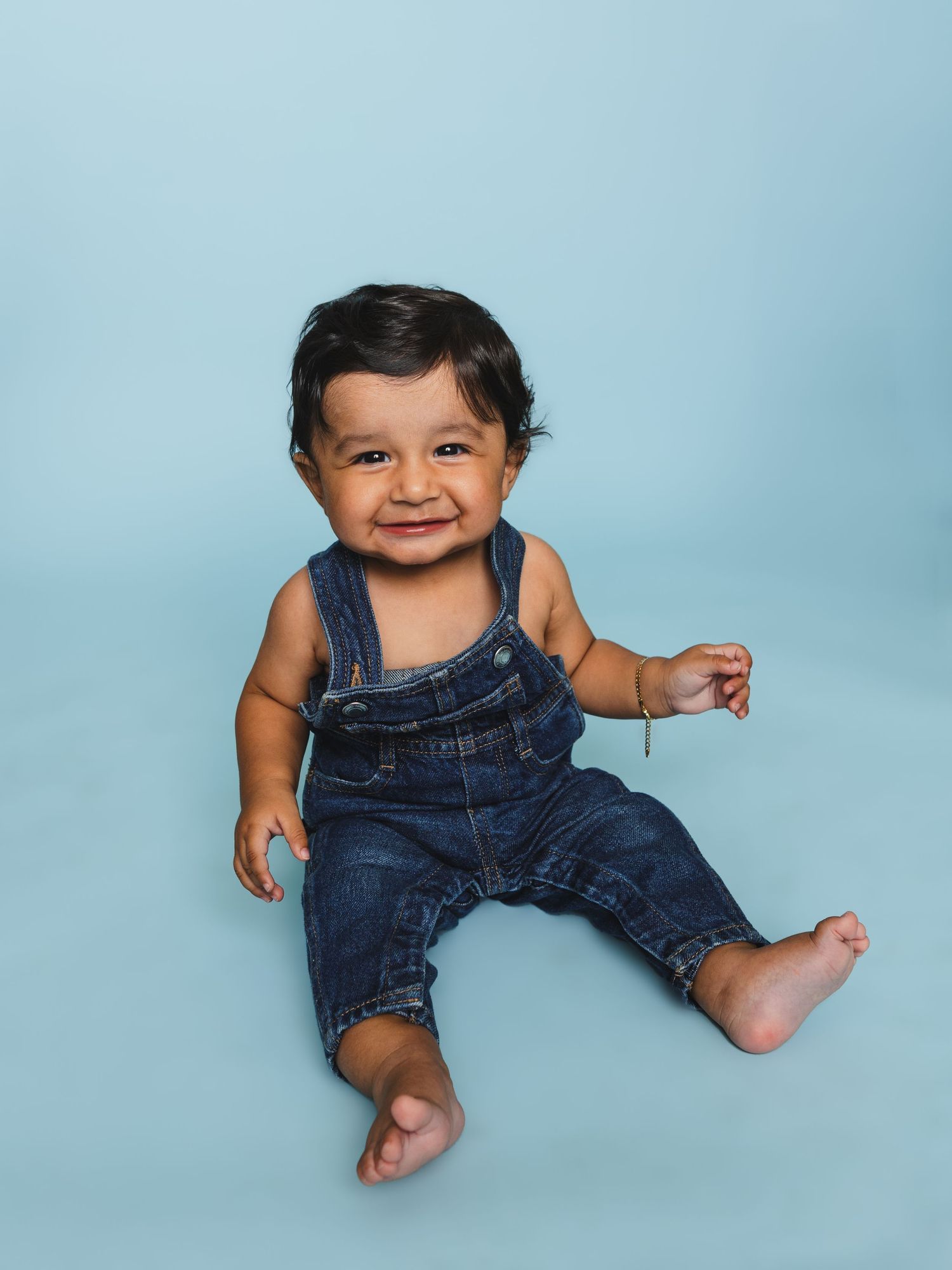 A happy baby wearing denim overalls sits against a light blue studio backdrop.