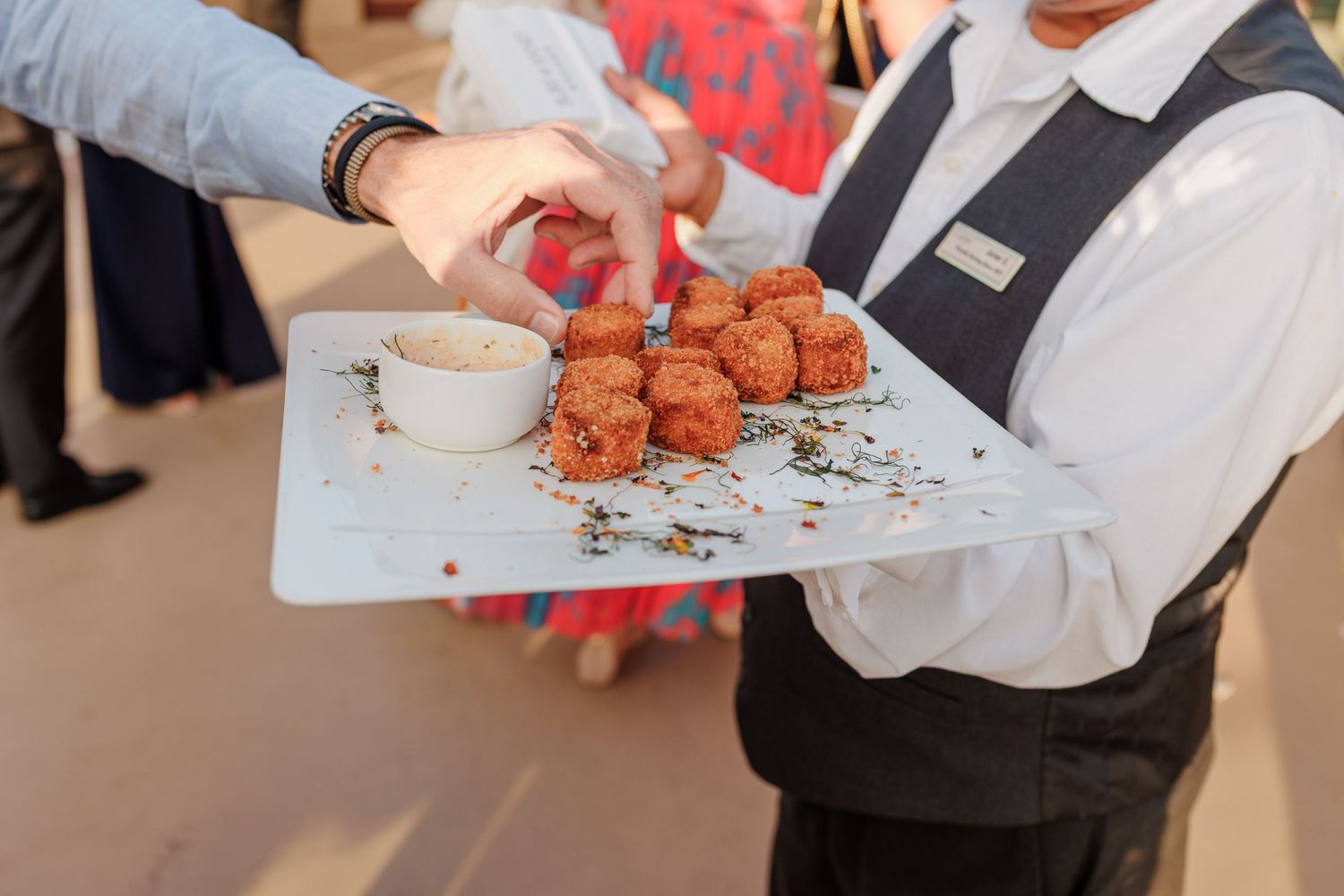 Waiter serving fried appetizers with dipping sauce on a white tray at a catered event.