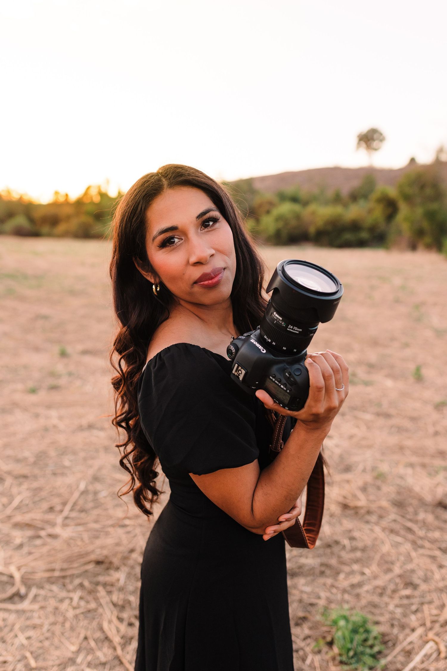 A photographer in a black dress poses with a professional camera in a desert landscape during golden hour.