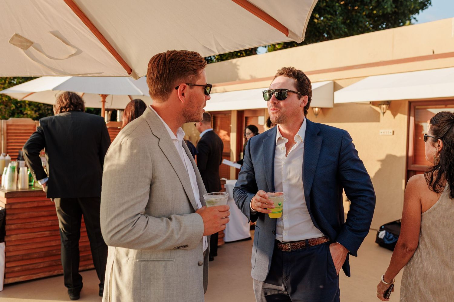 Two men in suits chat at an outdoor social event, holding drinks in a sunny venue.