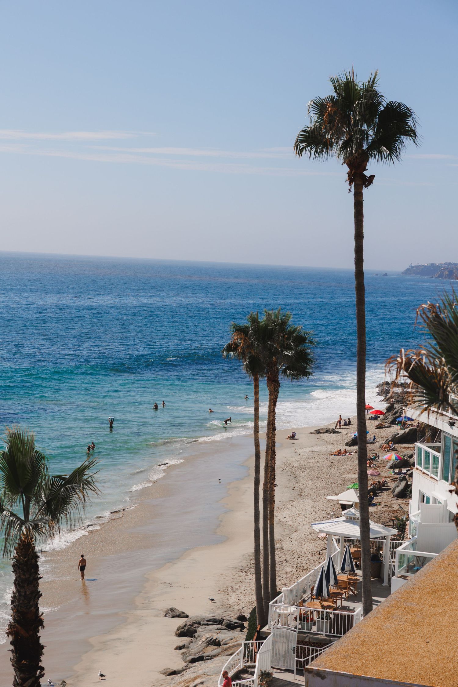 Aerial view of a sunny California Laguna beach with tall palm trees, turquoise waves, sunbathers, and beachfront properties.