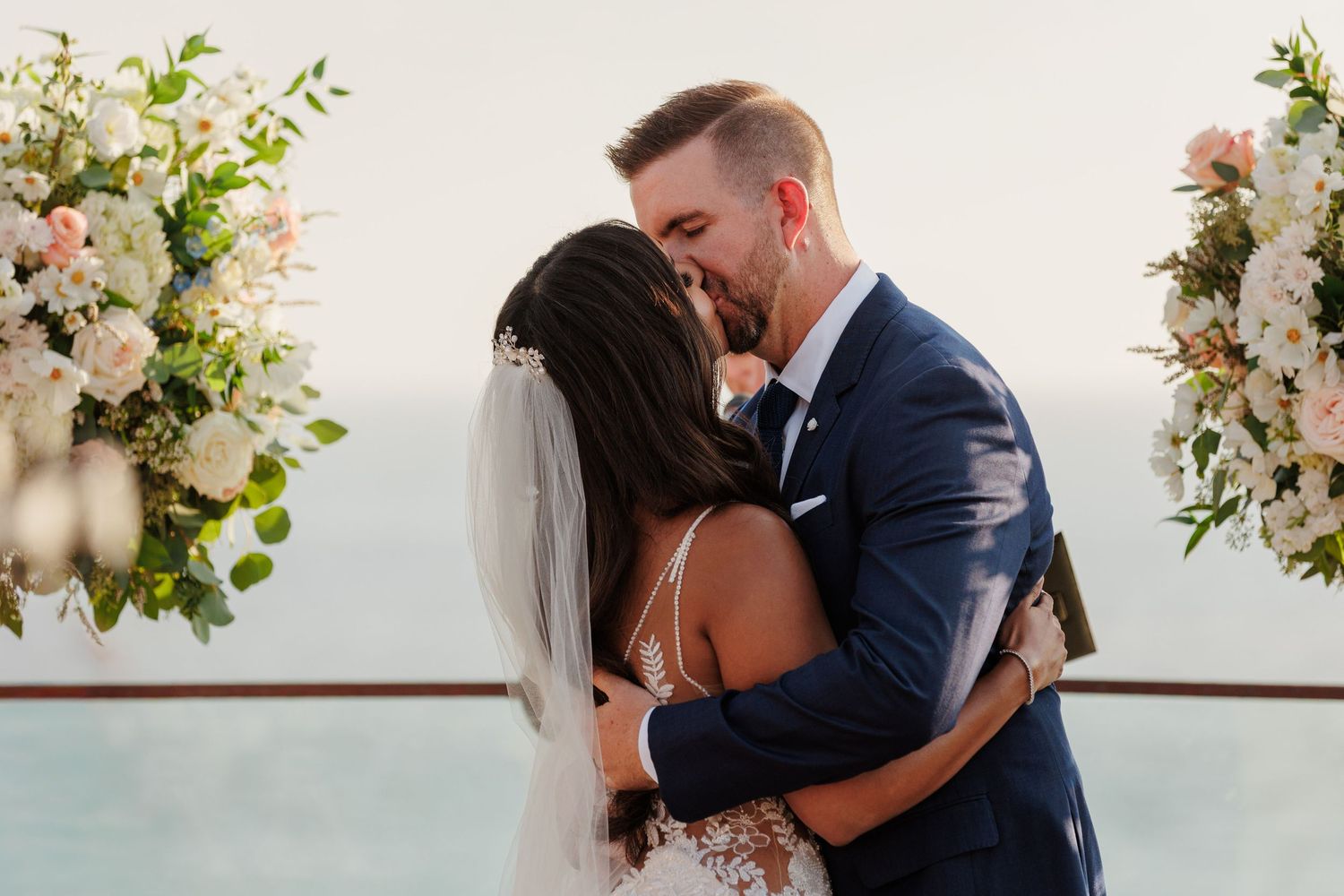Newlyweds share first kiss at outdoor wedding altar surrounded by white and pink floral arrangements.