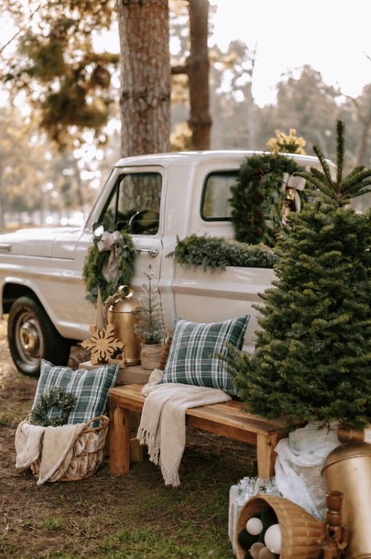 White vintage truck decorated with Christmas wreaths beside a wooden bench with plaid pillows and a small evergreen tree.