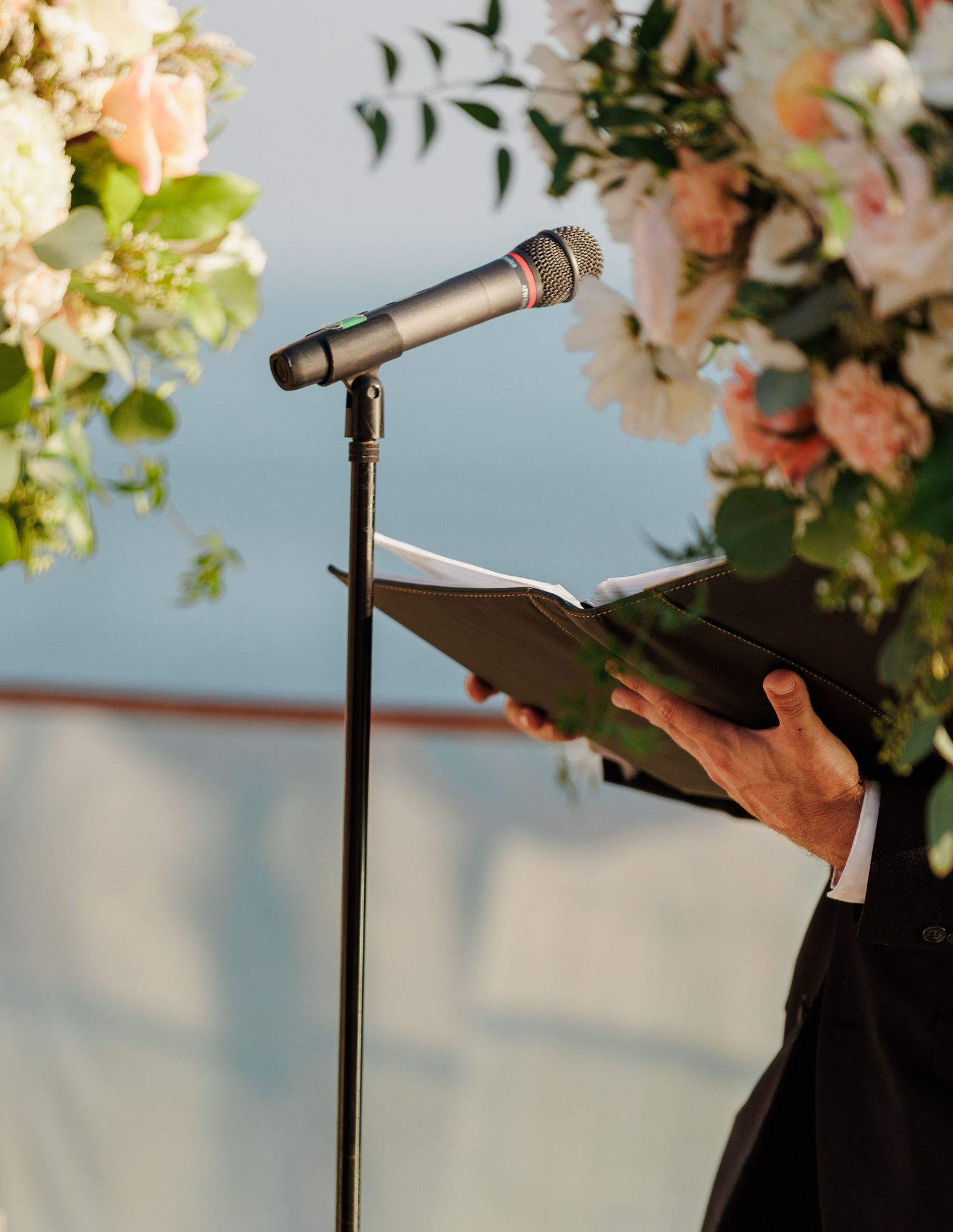 Officiant reads from a book at a wedding ceremony podium with a microphone and floral arrangement.