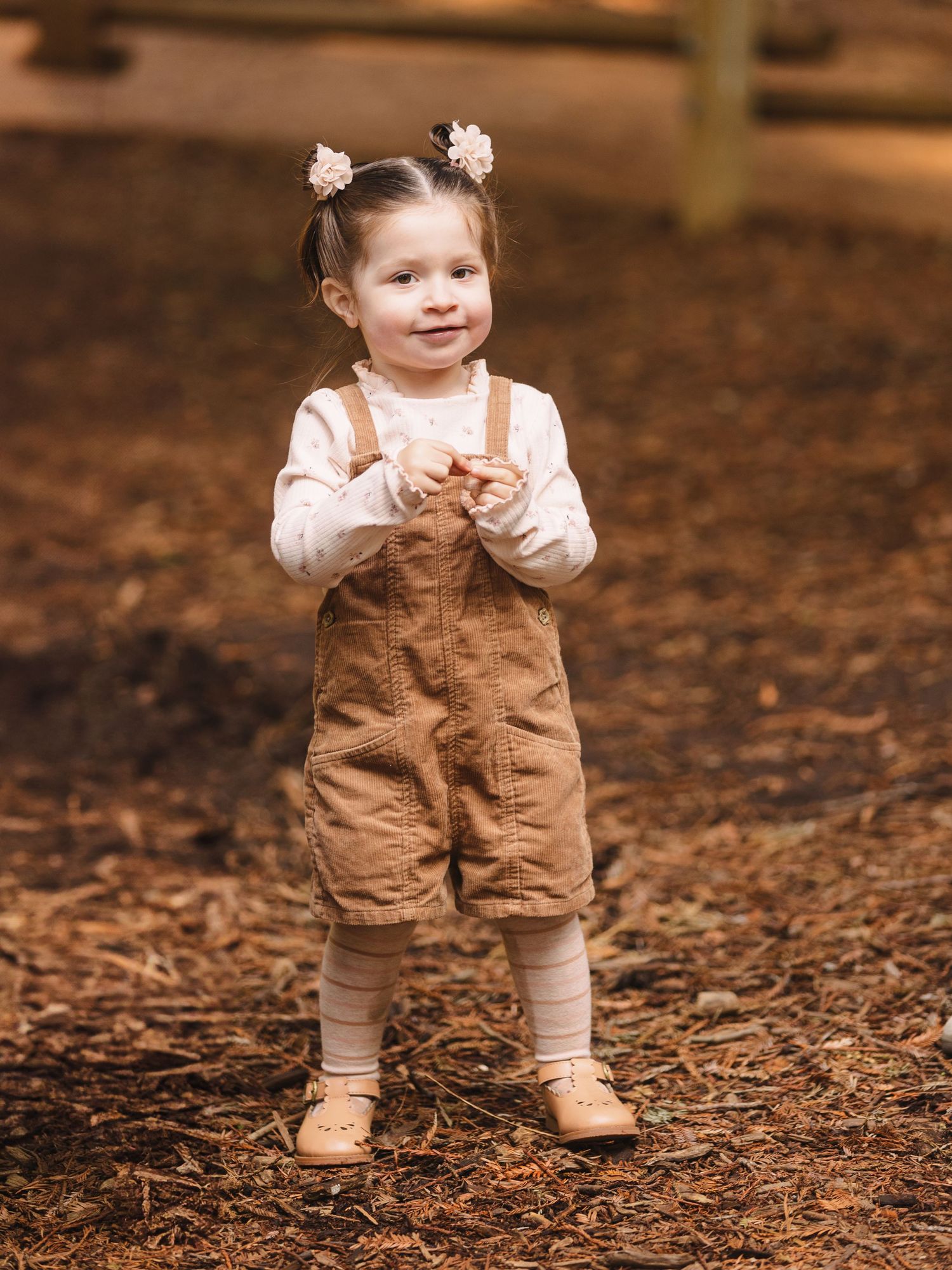 Toddler wearing brown overalls and white shirt stands on autumn leaves.